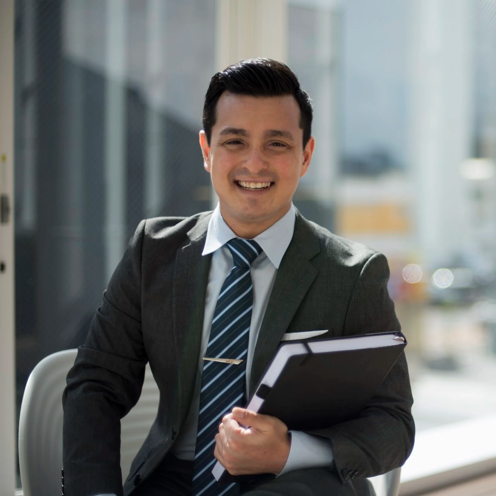 A smiling man in a business suit holding a folder, sitting in an office with large windows.