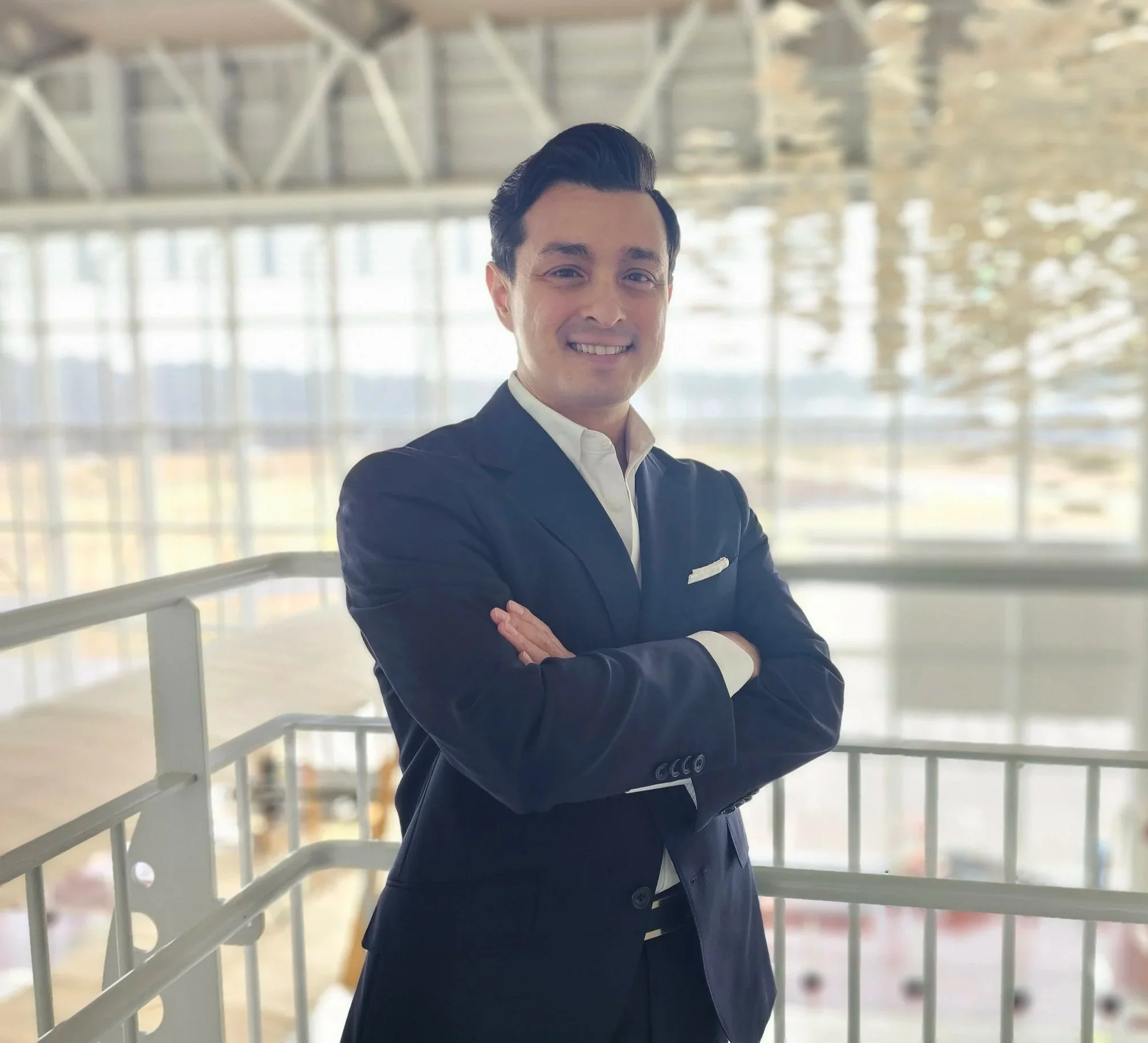 A man in a dark suit with a white shirt standing with arms crossed inside a modern building with large windows, smiling at the camera.