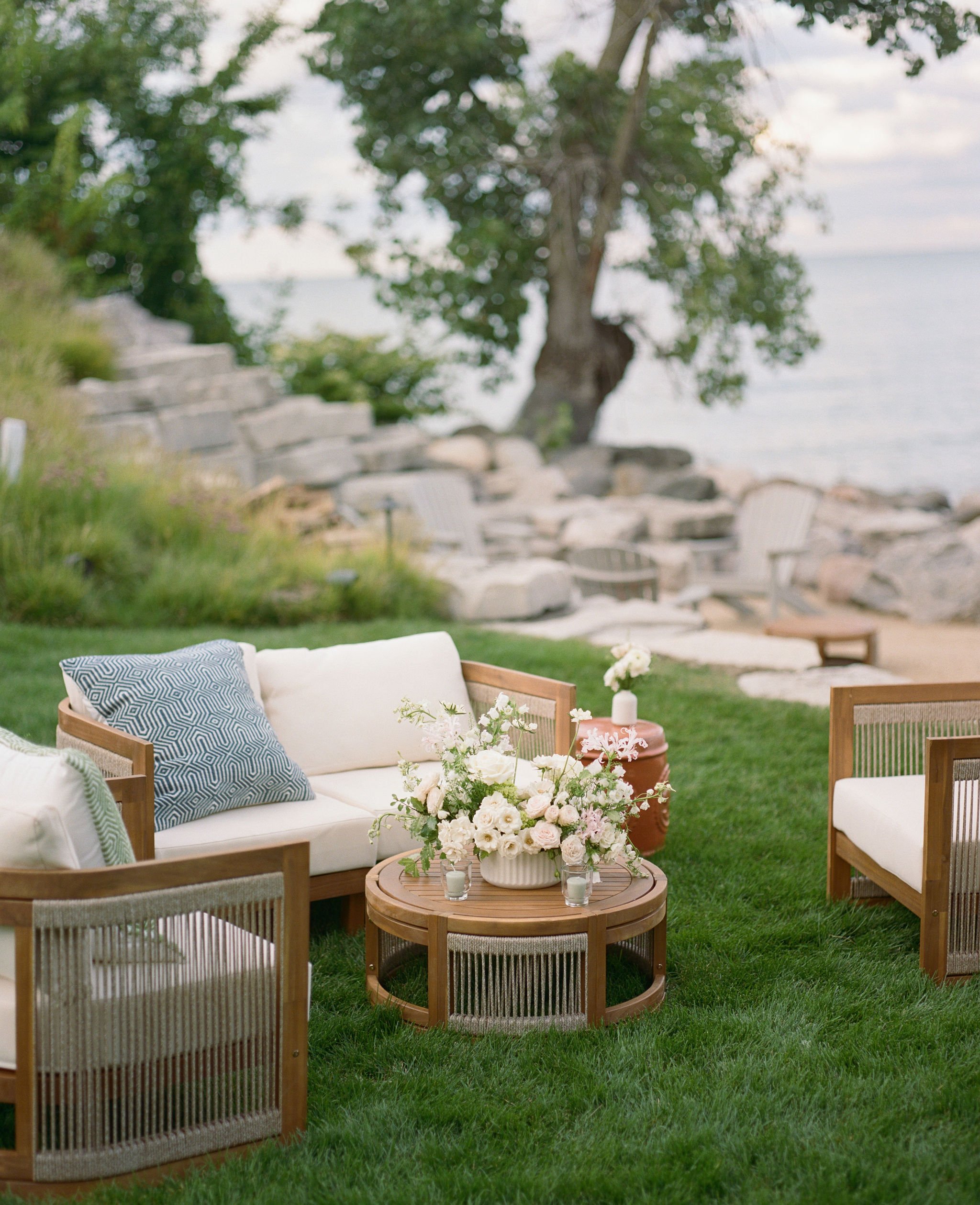 Outdoor patio area with cream-colored seating, floral arrangements on a round table, surrounded by green grass, trees, and views of a lake in the background.
