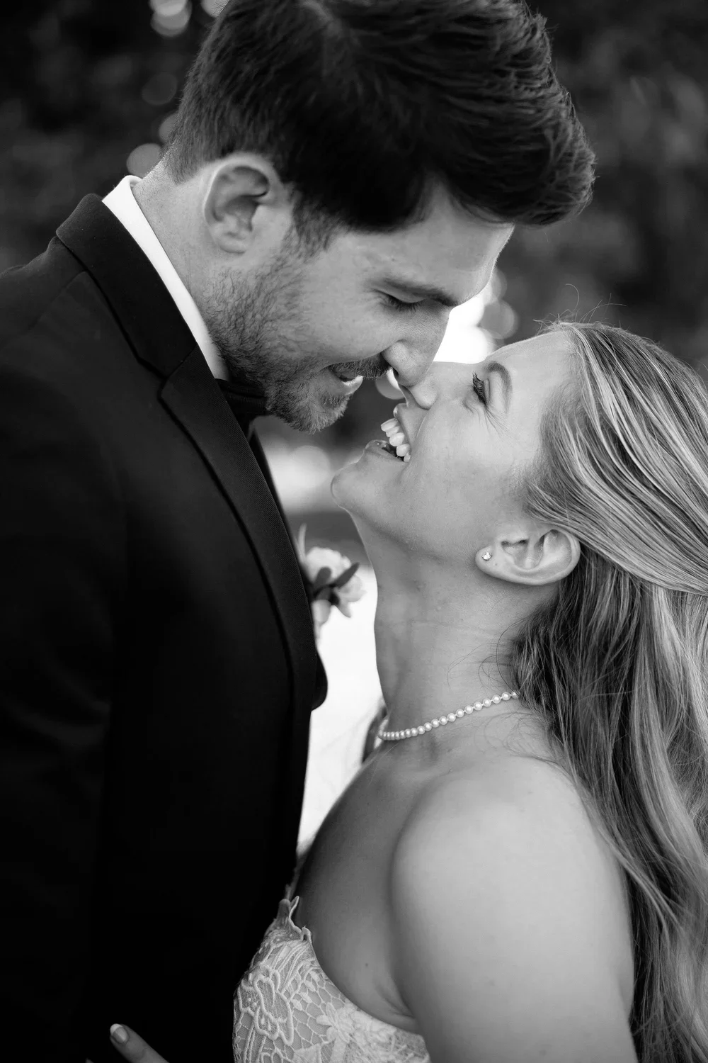 A black and white photo of a bride and groom smiling and leaning in close, about to kiss, outdoors.