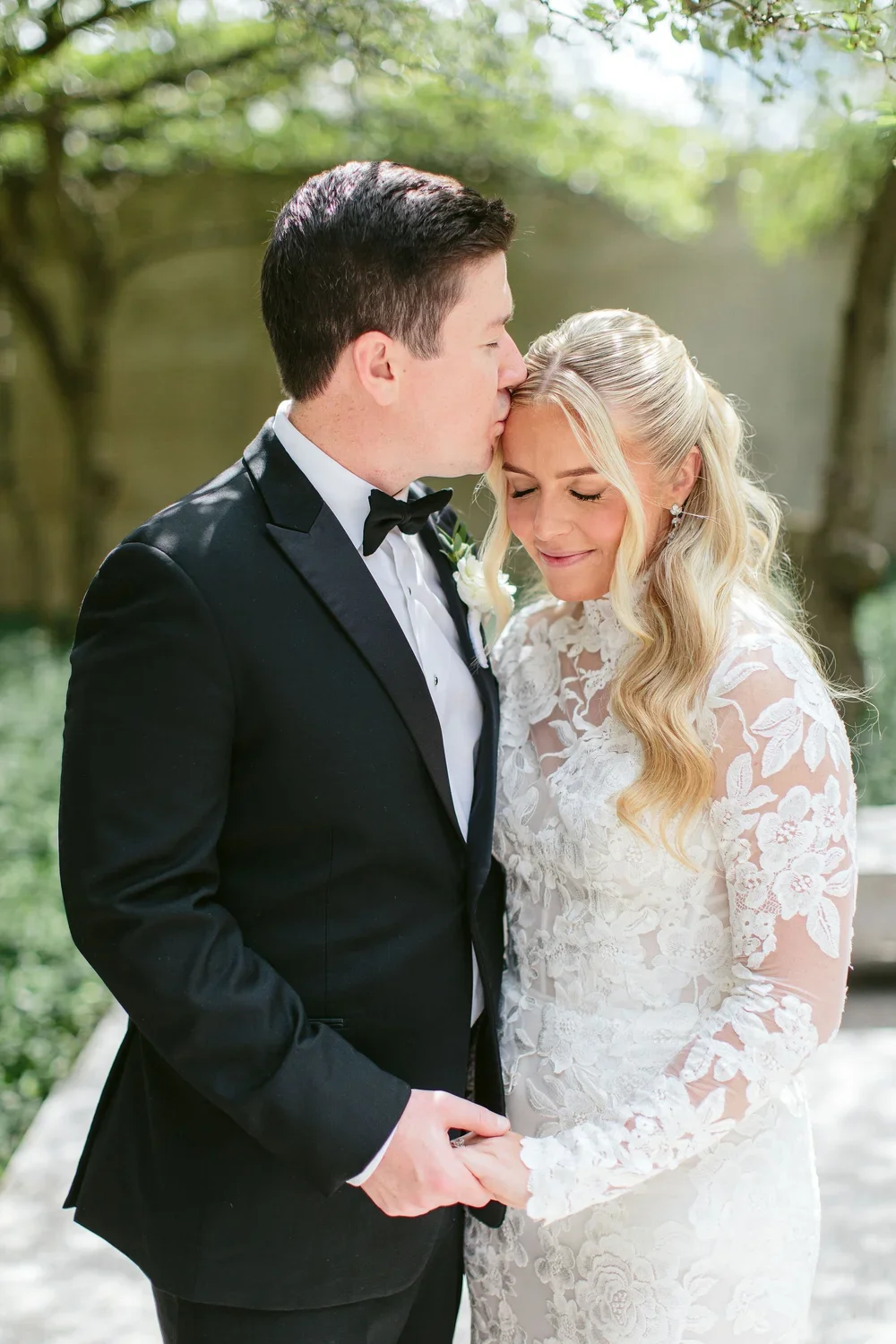 A groom in a black tuxedo kisses his bride on the forehead, holding her hands, during their wedding outside amidst greenery.