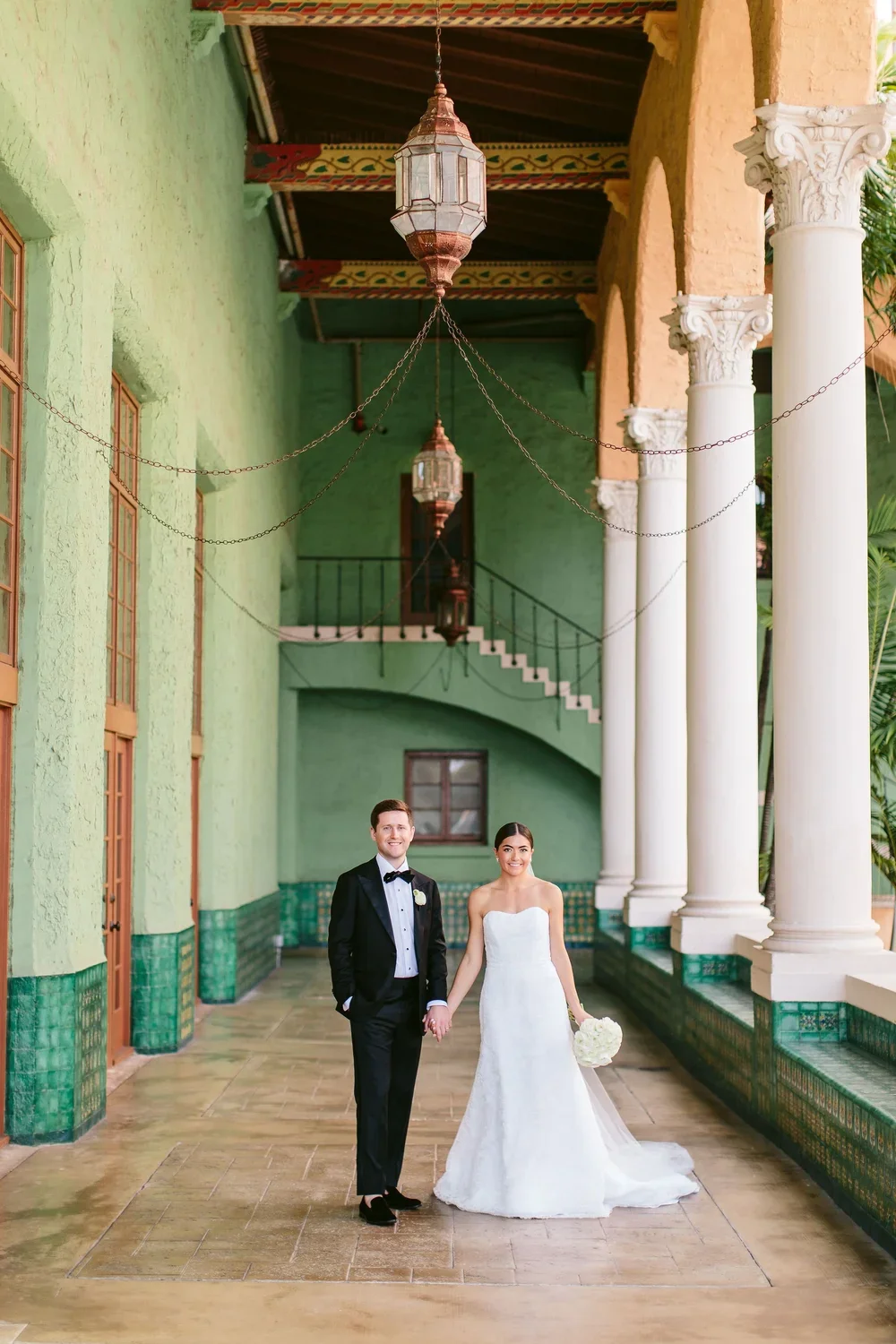 A bride and groom holding hands, standing in a colorful architectural corridor with tall white columns, green walls, and hanging lanterns, at a wedding celebration.