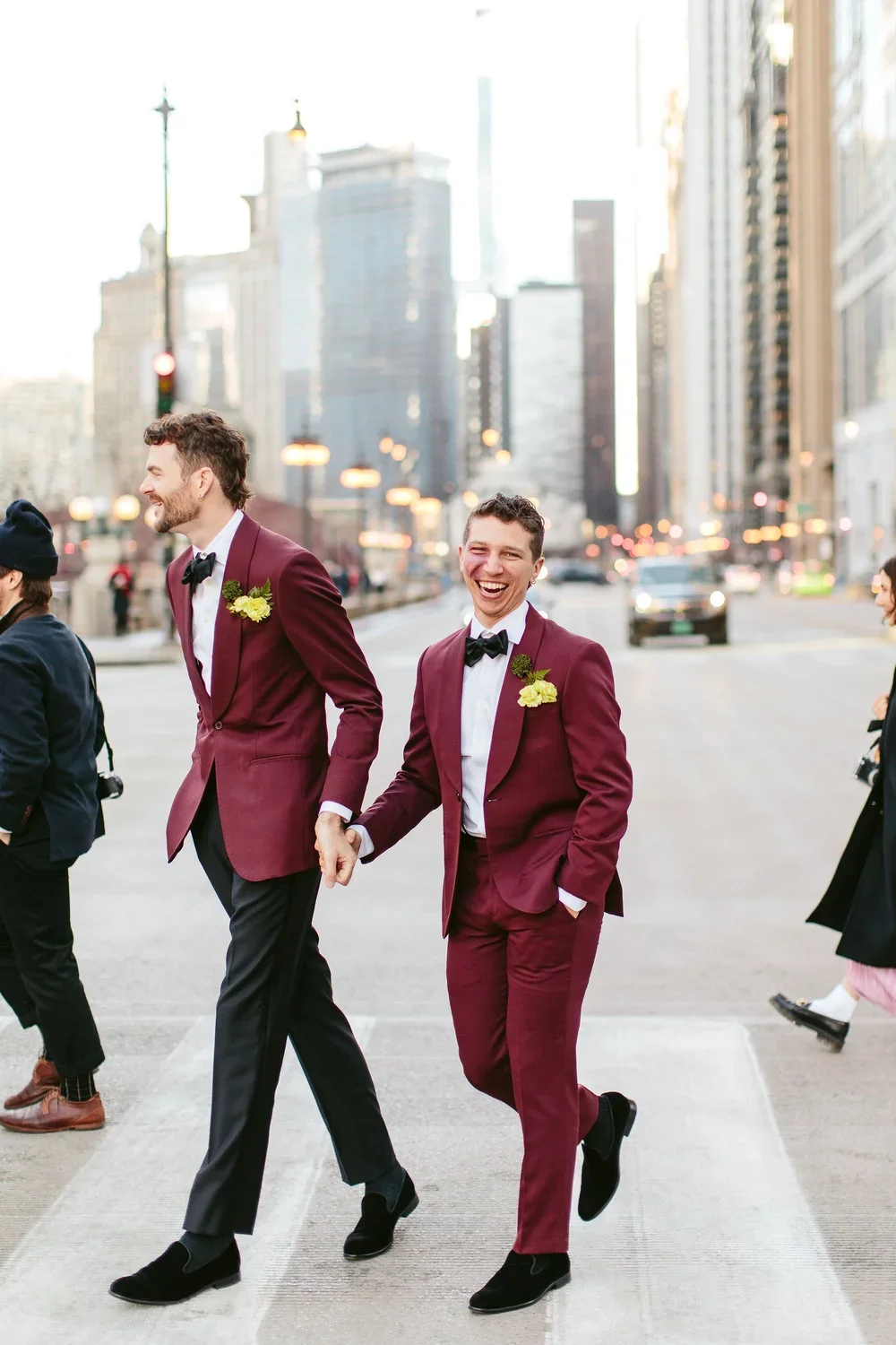 Two men in maroon suits with black bow ties and yellow boutonnières, holding hands and smiling while crossing a city street during daytime, with tall buildings and blurred traffic in the background.