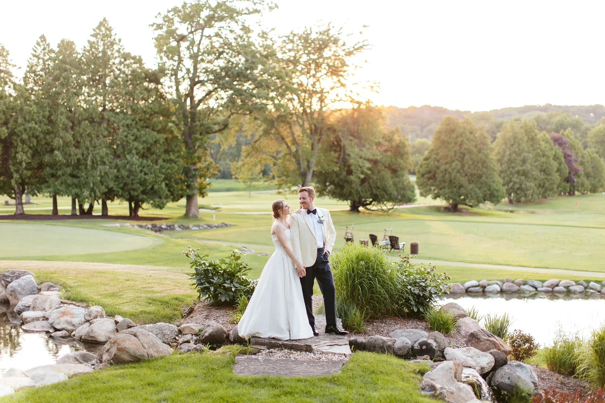 A bride and groom standing together outdoors by a pond, smiling at each other, with trees and a sunset in the background.