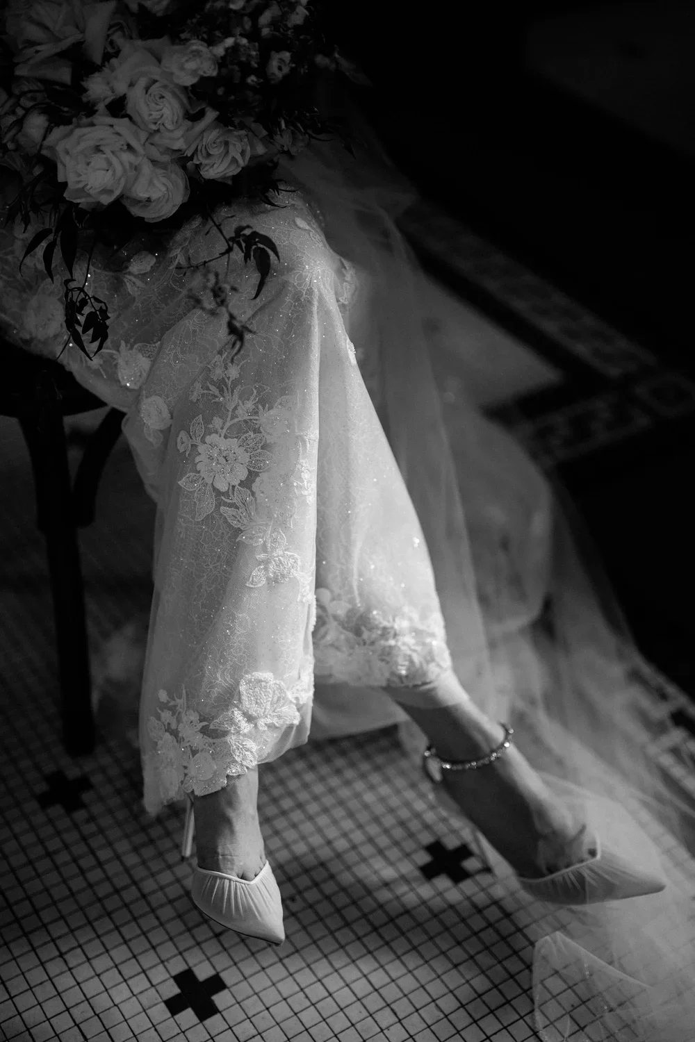 A woman sitting on a chair with legs crossed, wearing high heels and a long lace dress, holding a large bouquet of roses.