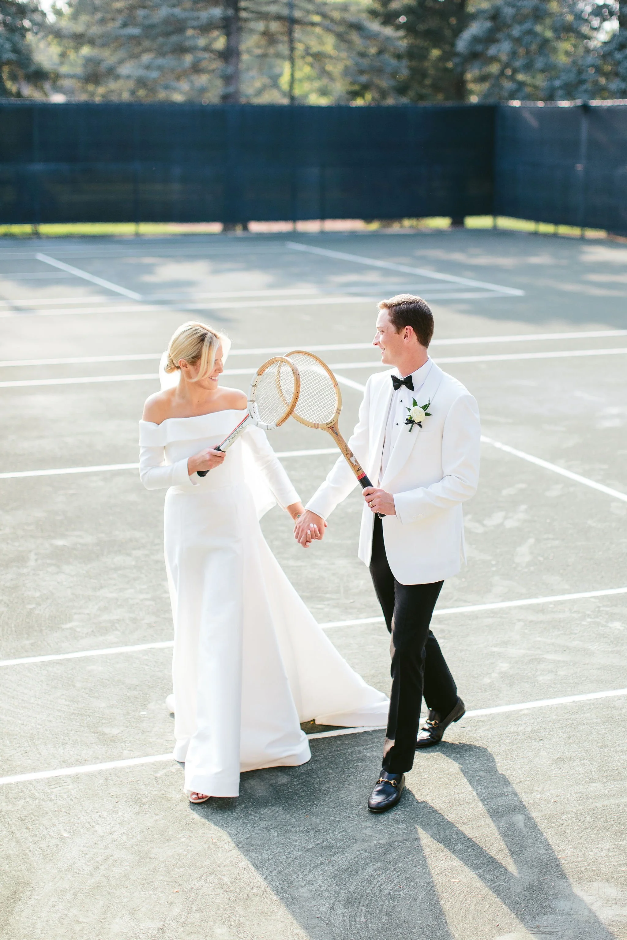 A bride and groom in wedding attire are standing on a tennis court, holding tennis rackets, and smiling at each other, with one a woman in a white wedding dress and the other a man in a white tuxedo jacket with a black bow tie.