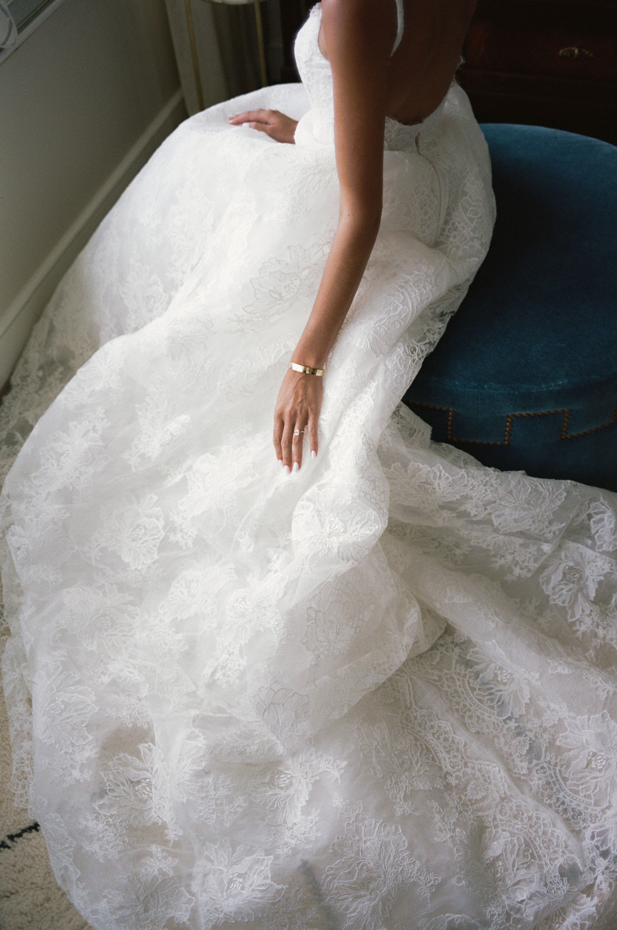 A woman in a white lace wedding dress with floral patterns, sitting on a blue velvet chair, showing her left arm with a gold bracelet and a ring, in a room with beige carpet.
