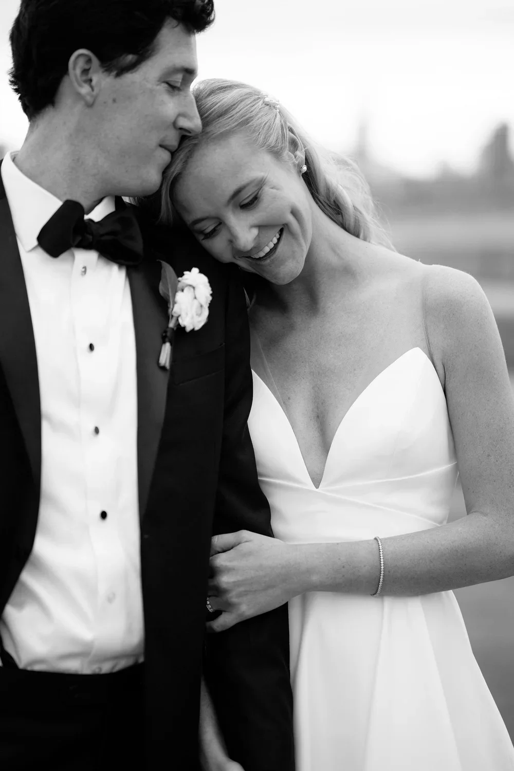 A black-and-white photo of a bride and groom sharing a tender moment, smiling and leaning their heads together outdoors.