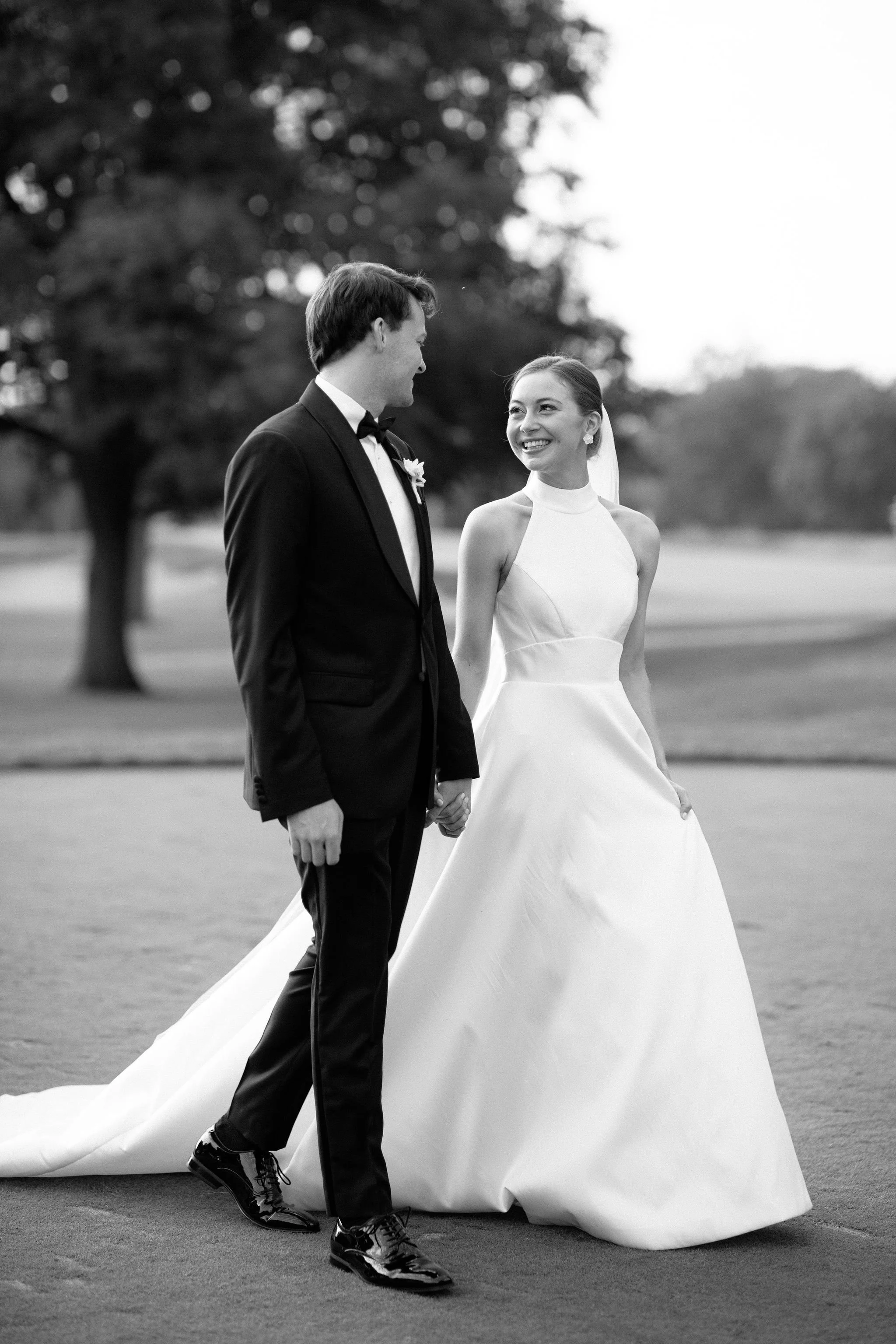 A black and white photo of a smiling couple in wedding attire, holding hands outdoors on a grassy area with trees in the background. The man is wearing a tuxedo and the woman is in a wedding dress with a veil.