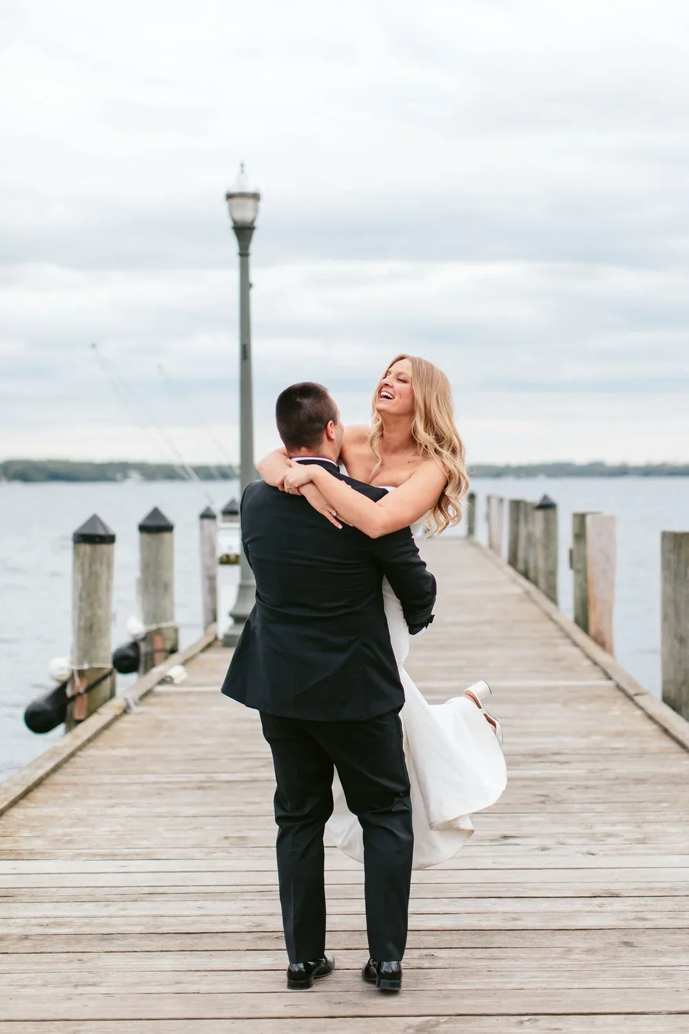 A man in a black suit lifts a smiling woman in a white dress on a pier overlooking water and cloudy sky.