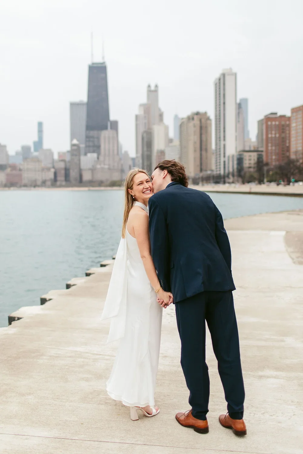A couple holding hands and sharing a kiss on a lakeside walkway with the Chicago skyline in the background.