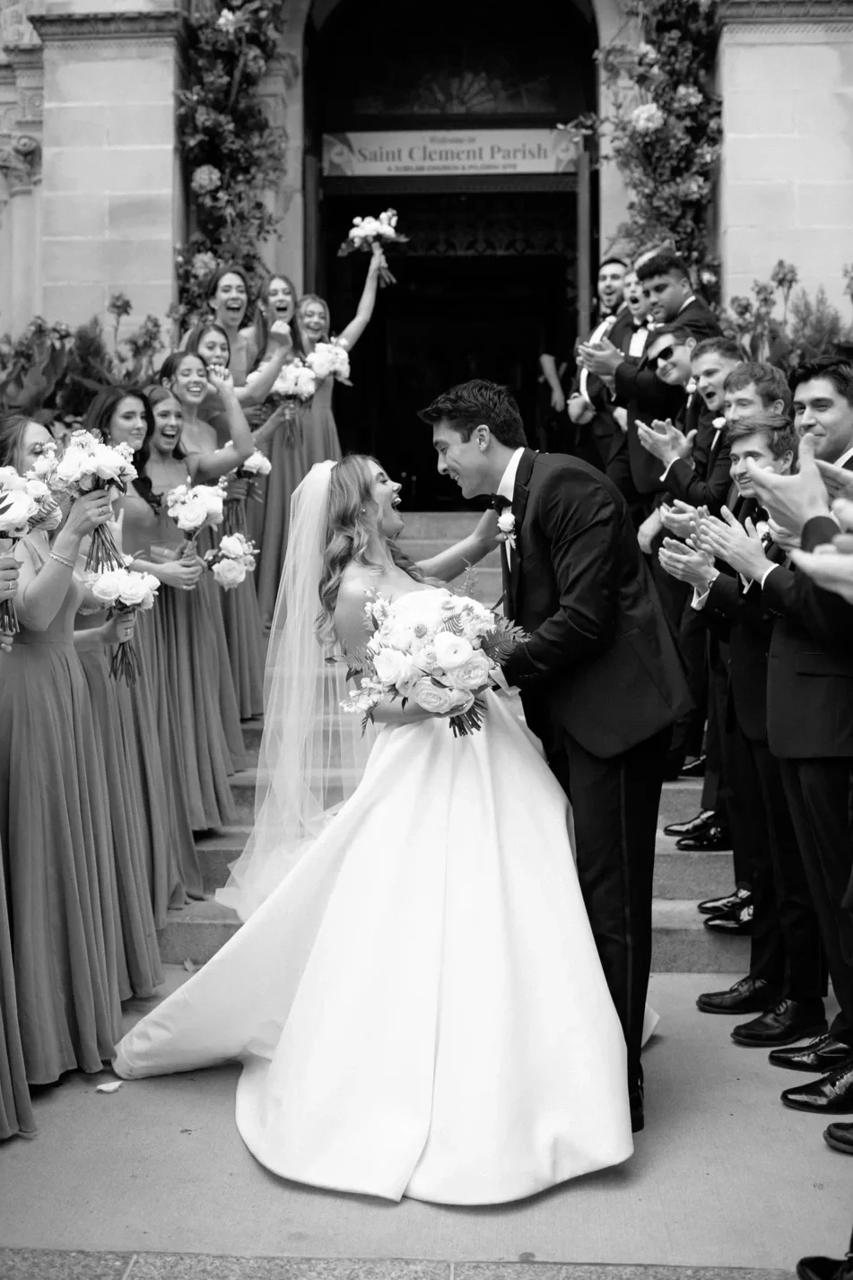 A bride and groom embrace in front of a large group of wedding guests, outside a church decorated with flowers, as the bride holds a bouquet and they smile at each other.