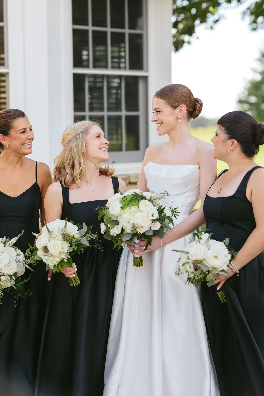 A bride in a white wedding gown and four bridesmaids in black dresses holding bouquets of white flowers outside a house with green trees in the background.