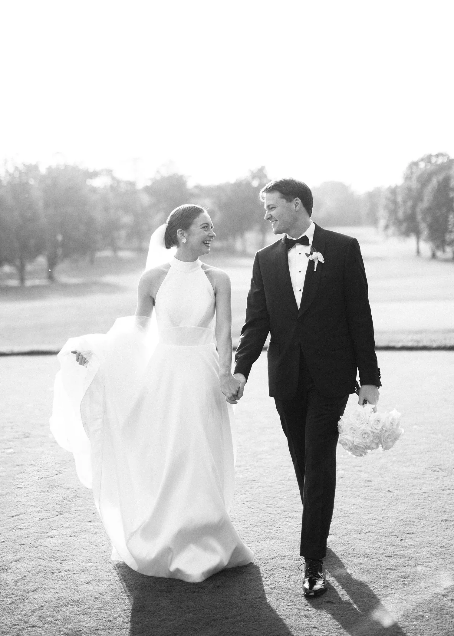 A black and white photo of a bride and groom holding hands, smiling at each other outdoors on a sunny day. The bride is in a white wedding dress with a veil, and the groom is in a tuxedo holding a bouquet of white roses.