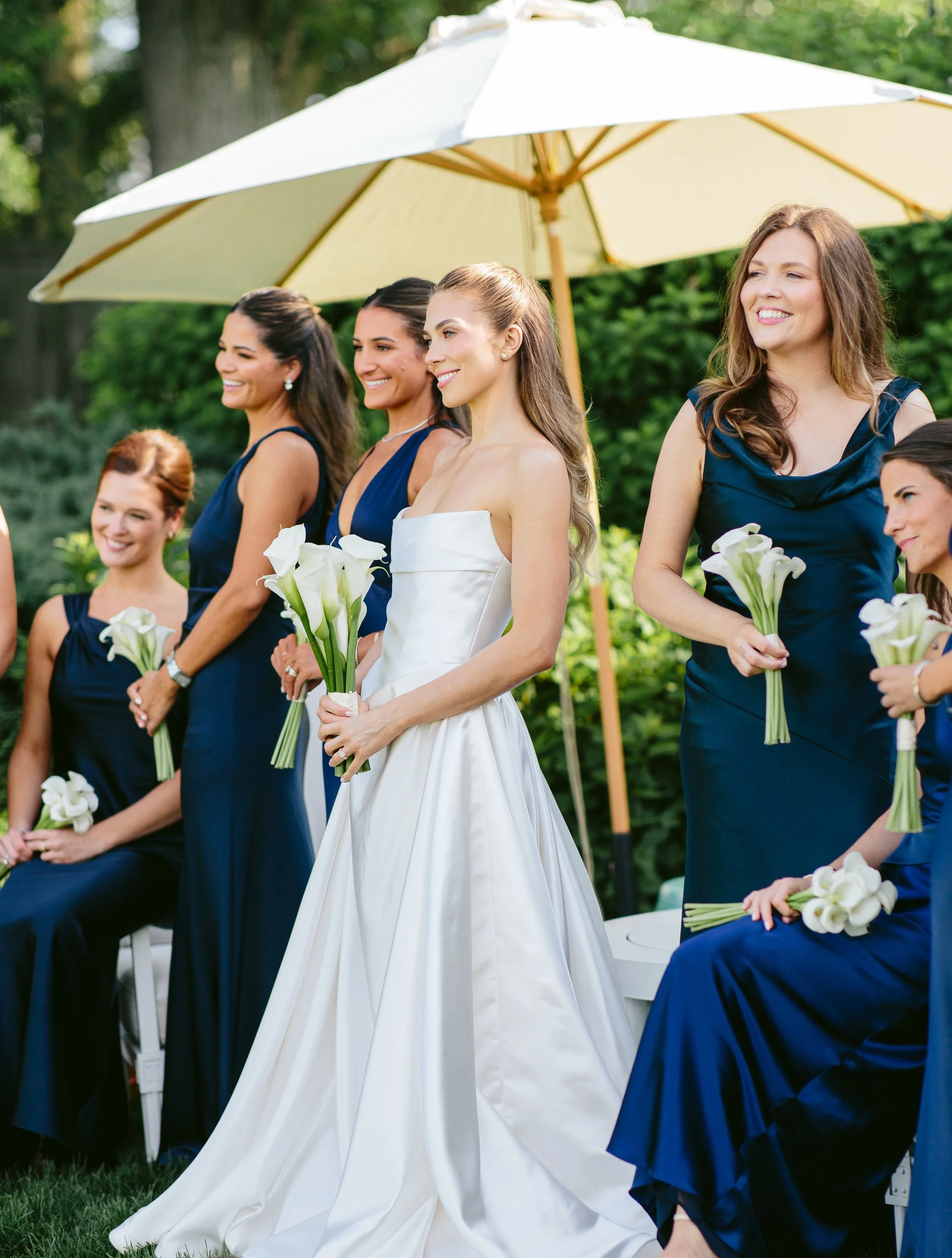 Bride in white gown holding calla lilies, surrounded by women in navy dresses at outdoor wedding ceremony with greenery and large white umbrella in background.