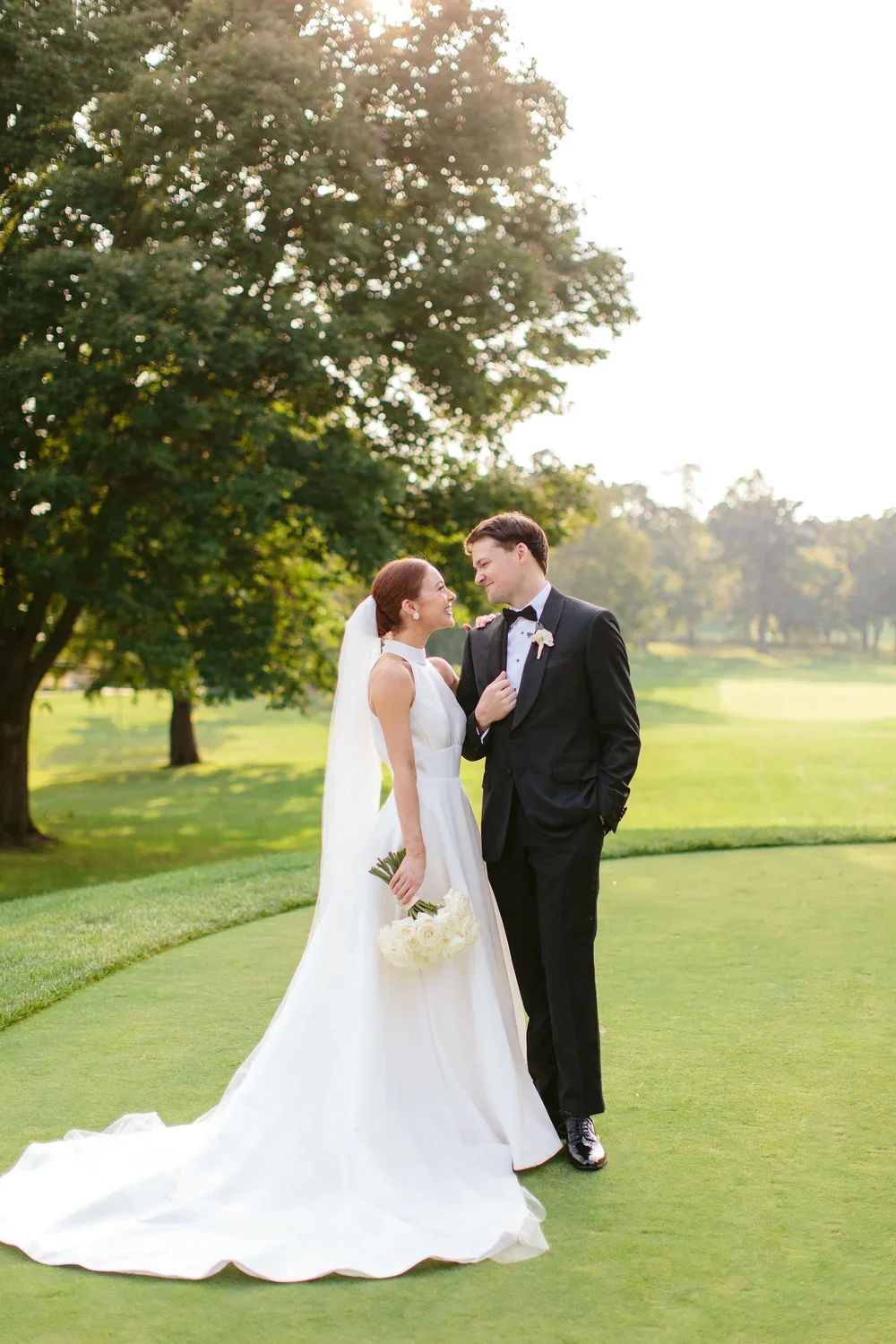 A bride and groom standing on a lush green golf course, gazing at each other, with the bride holding a bouquet of white flowers and the groom in a tuxedo, surrounded by trees under a bright sky.