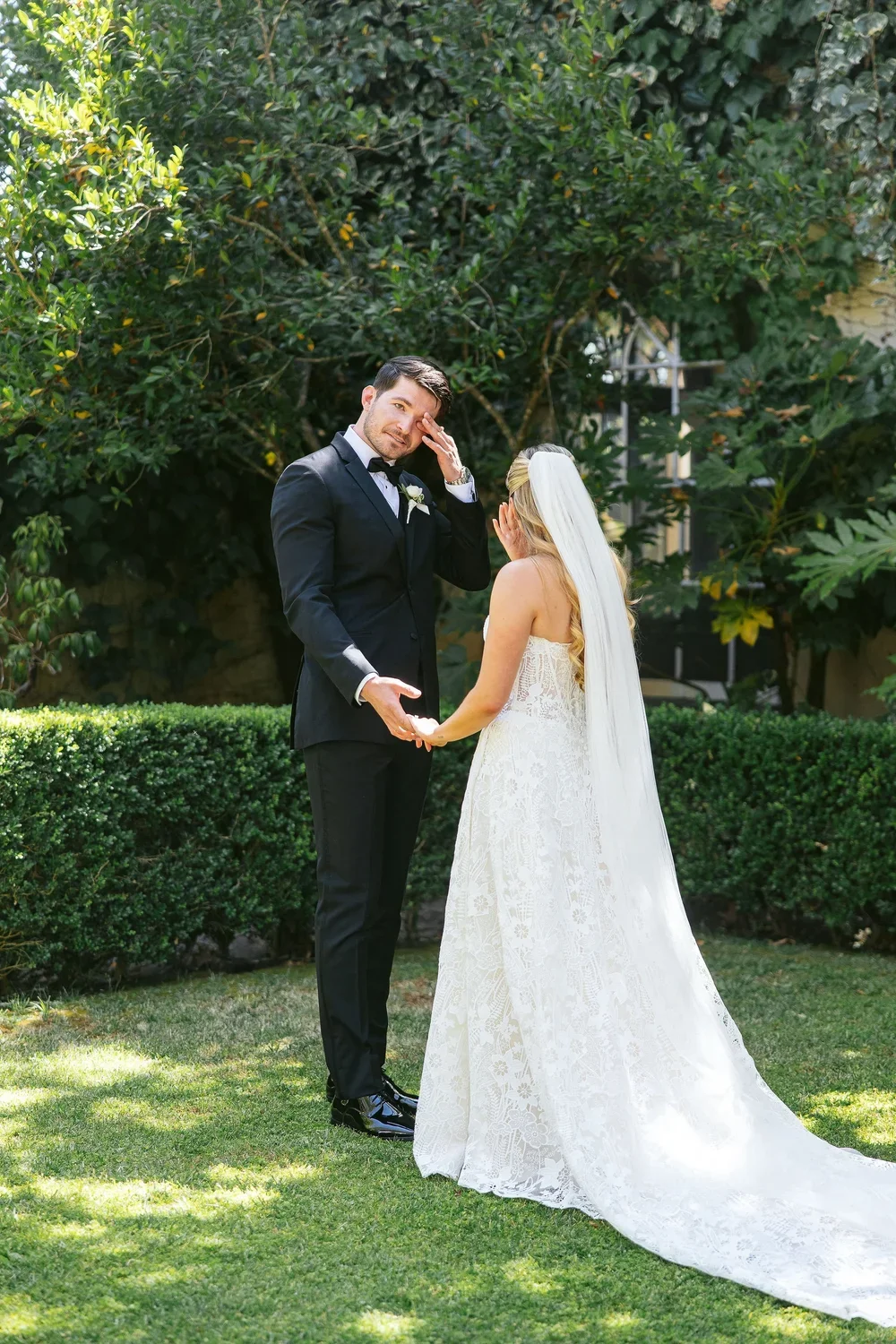 A bride and groom stand outdoors, holding hands, with the groom appearing emotional. The bride is wearing a white lace wedding gown and a long veil. The groom is in a black tuxedo with a boutonniere. They are surrounded by greenery, trees, and a manicured lawn.