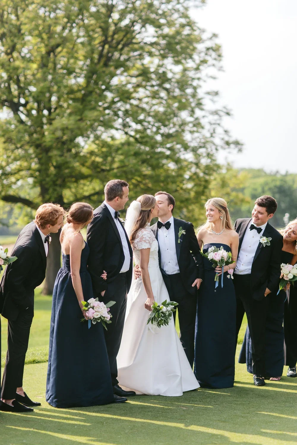 A wedding party outdoors, with the bride and groom kissing, surrounded by friends holding bouquets, on a sunny day with green trees in the background.