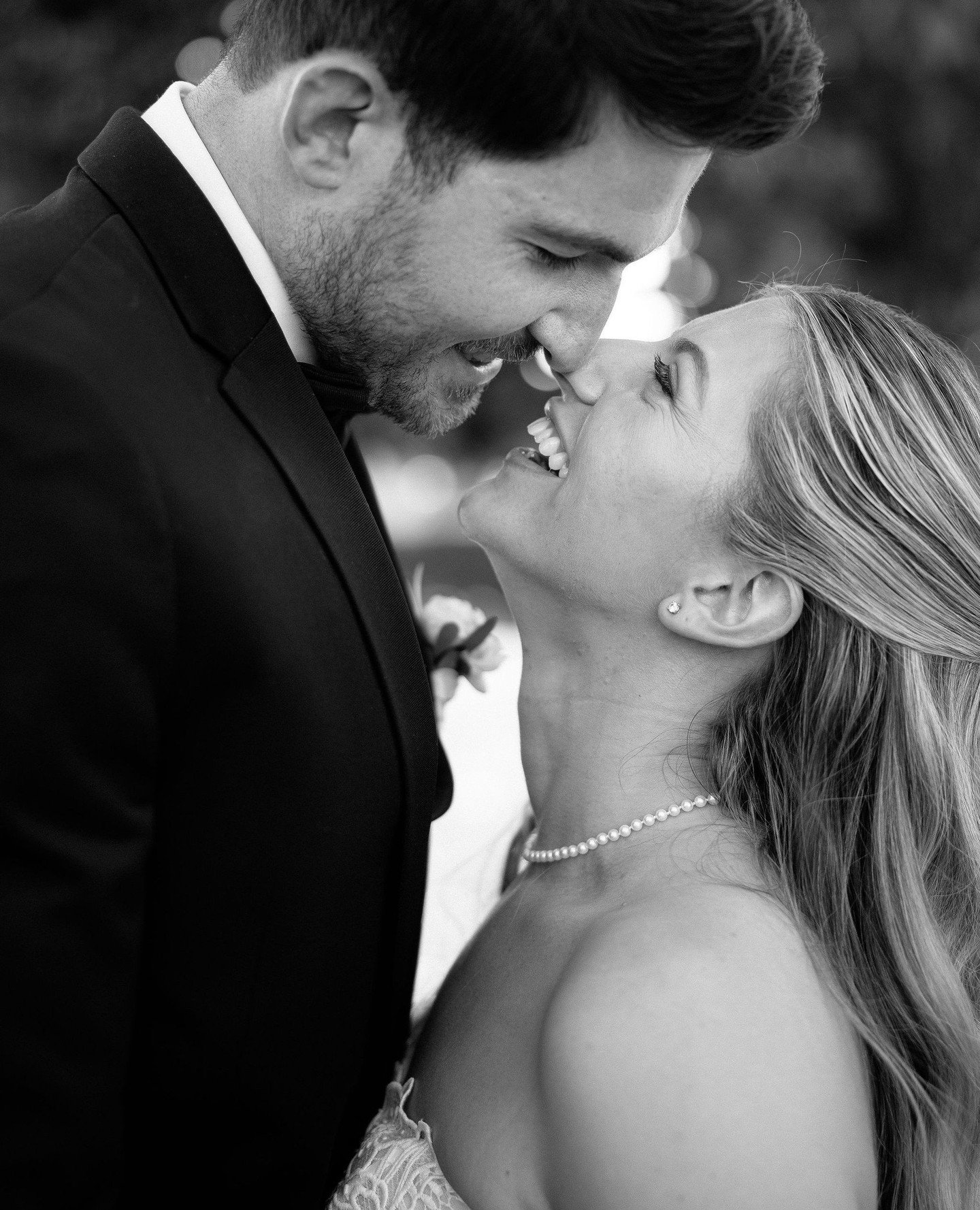 Black and white photo of a smiling woman with long hair wearing a pearl necklace, looking up at a man with a beard, wearing a tuxedo, who is leaning down towards her with their noses touching.