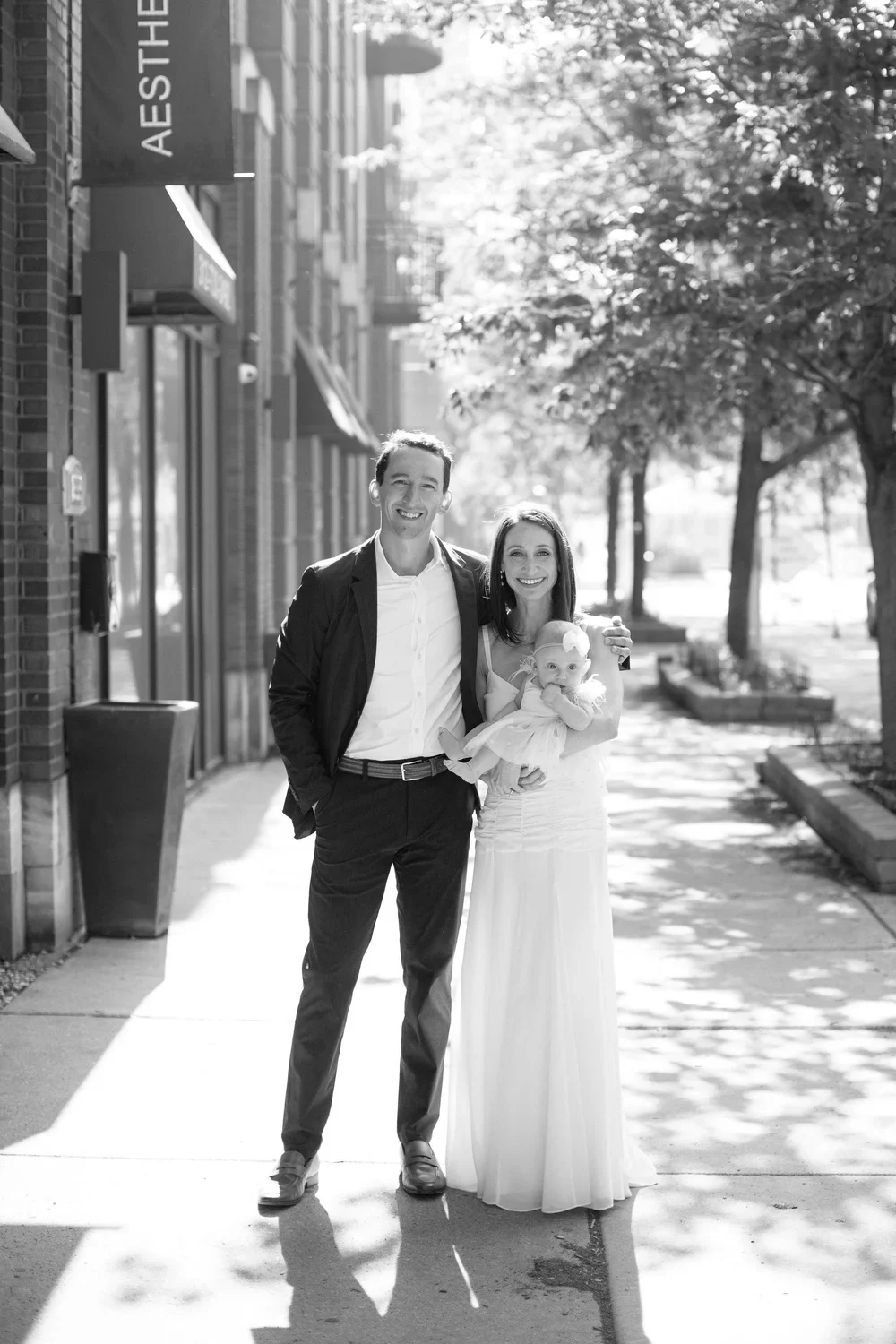 A happy family of three, including a man, woman, and a baby girl in a white dress, standing on a sidewalk in an urban area with trees and buildings in the background, sunlight casting shadows.