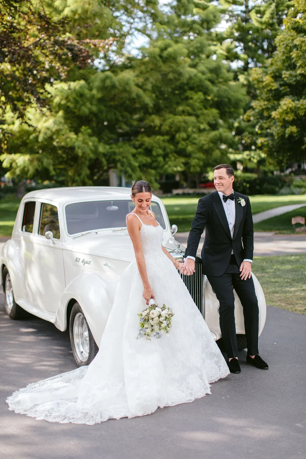 Bride and groom in wedding attire holding hands next to a vintage white car in a park with green trees.