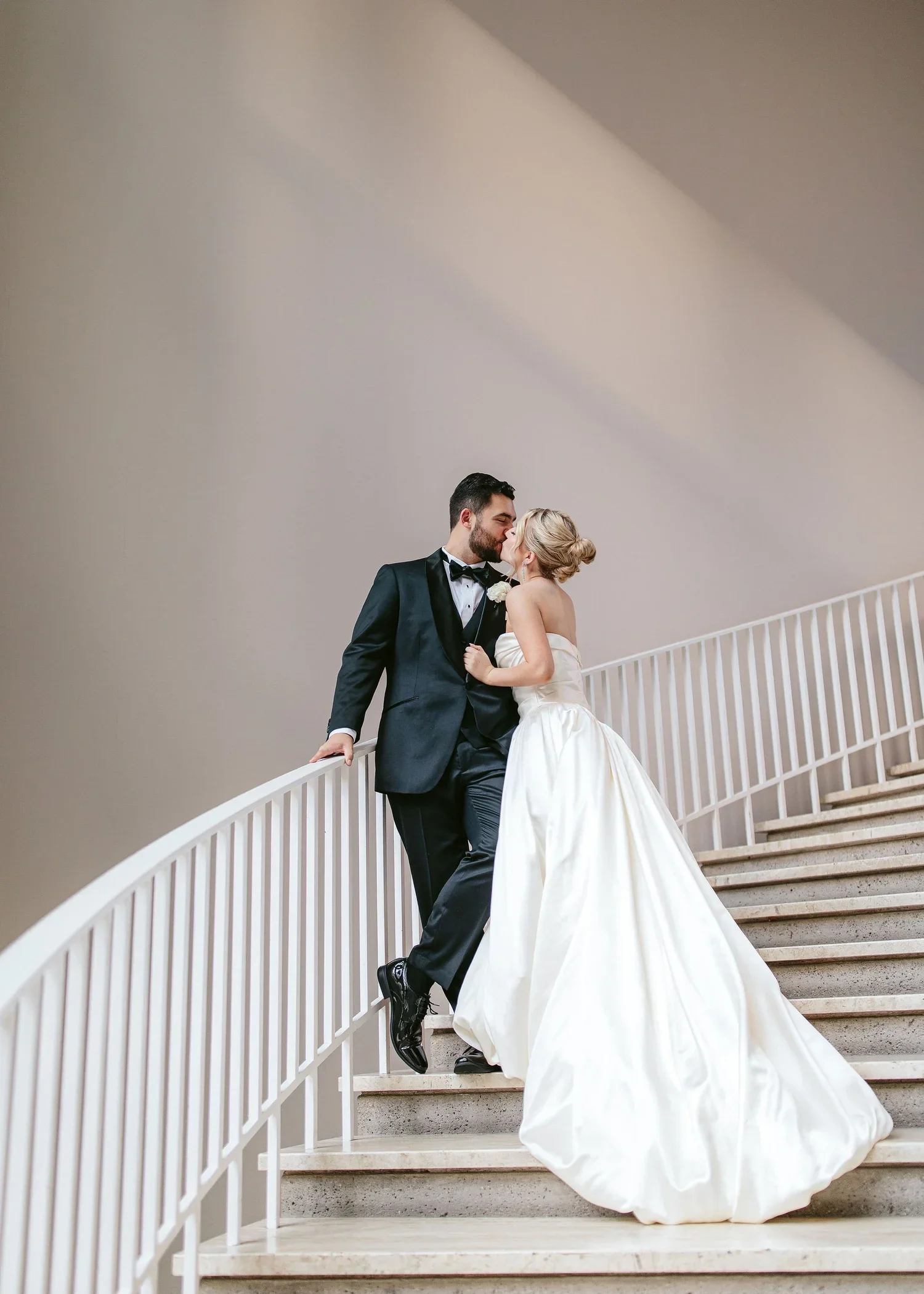 A bride and groom are sharing a kiss on a staircase. The groom is dressed in a black tuxedo and the bride in a strapless white wedding gown. The staircase has a white railing and the background features a plain wall with a diagonal shadow.