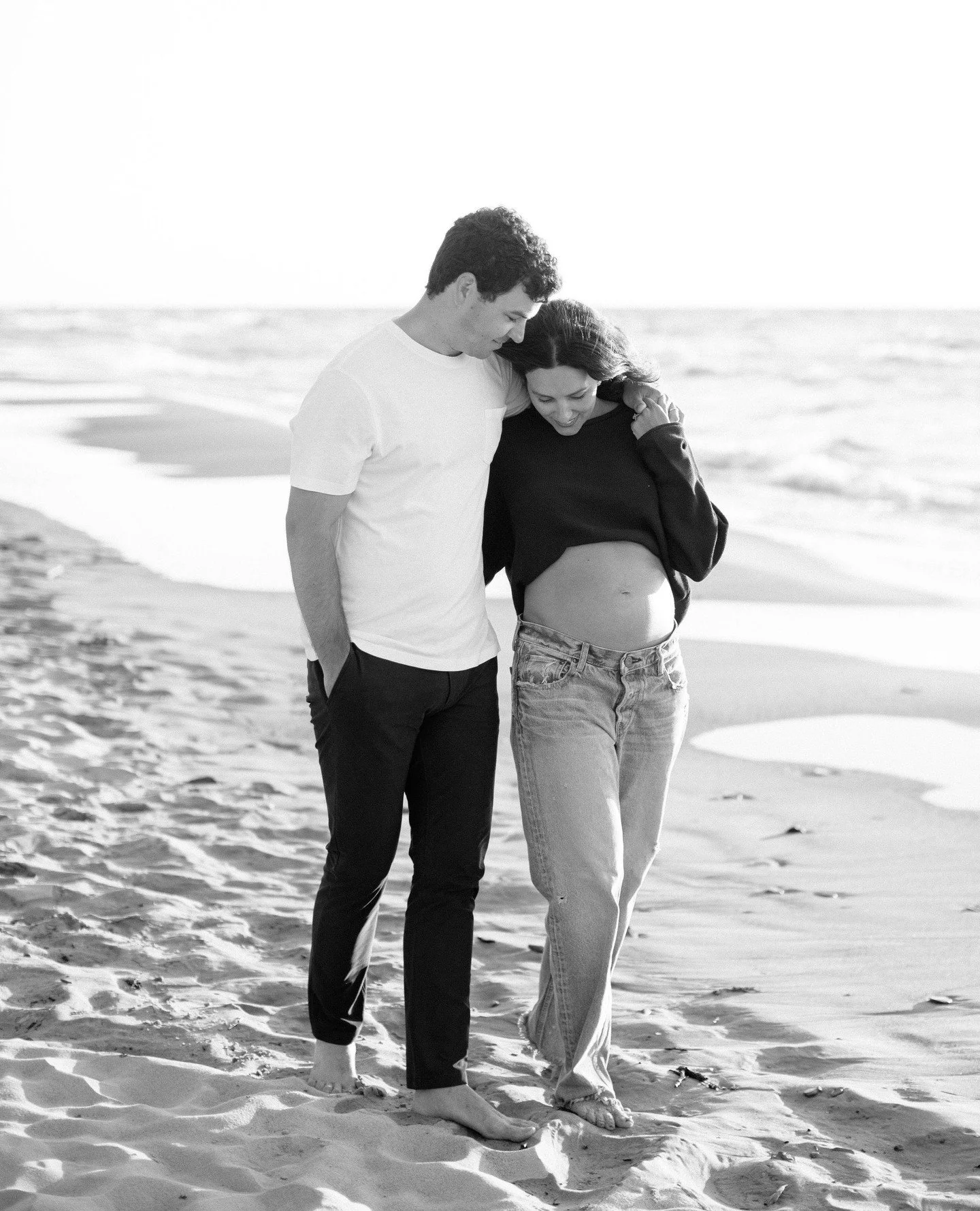 A couple walking along the beach, smiling and looking at a pregnant woman's belly, while holding hands and leaning their foreheads together.