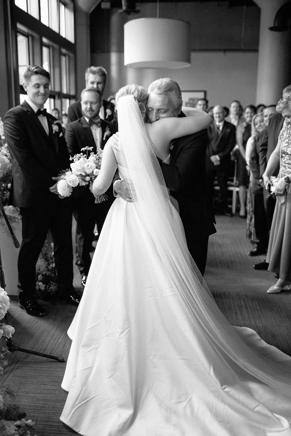 A bride and an older man sharing an emotional hug during a wedding ceremony, with guests watching and smiling in the background.