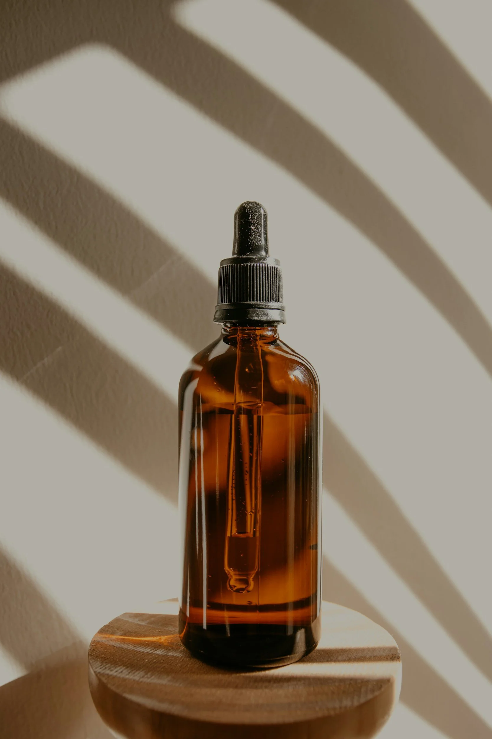 A brown glass dropper bottle with a black cap and dropper inside, placed on a wooden surface with sunlight casting striped shadows on a light-colored wall.