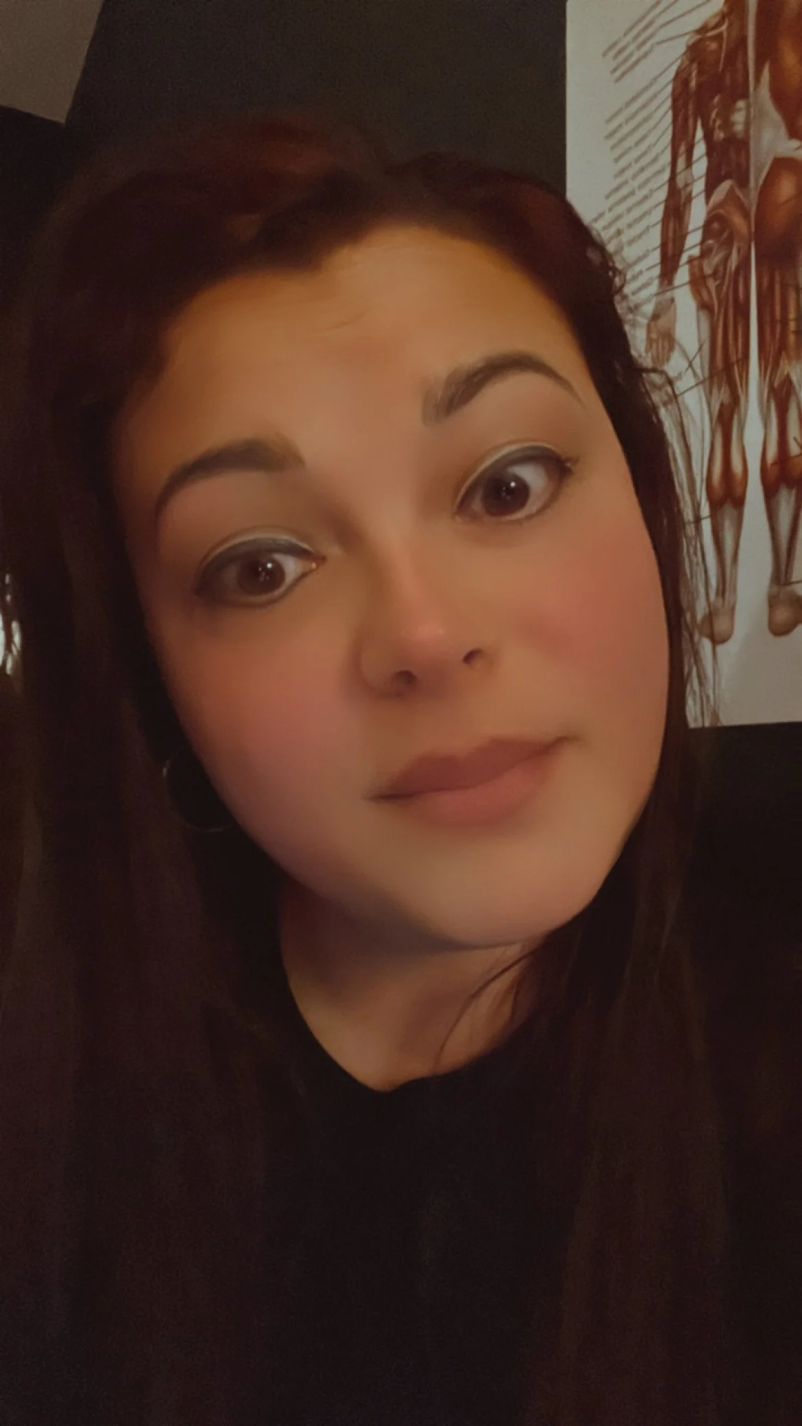 Close-up of a woman's face with makeup, dark hair, and earrings, sitting in front of a medical diagram on the wall.