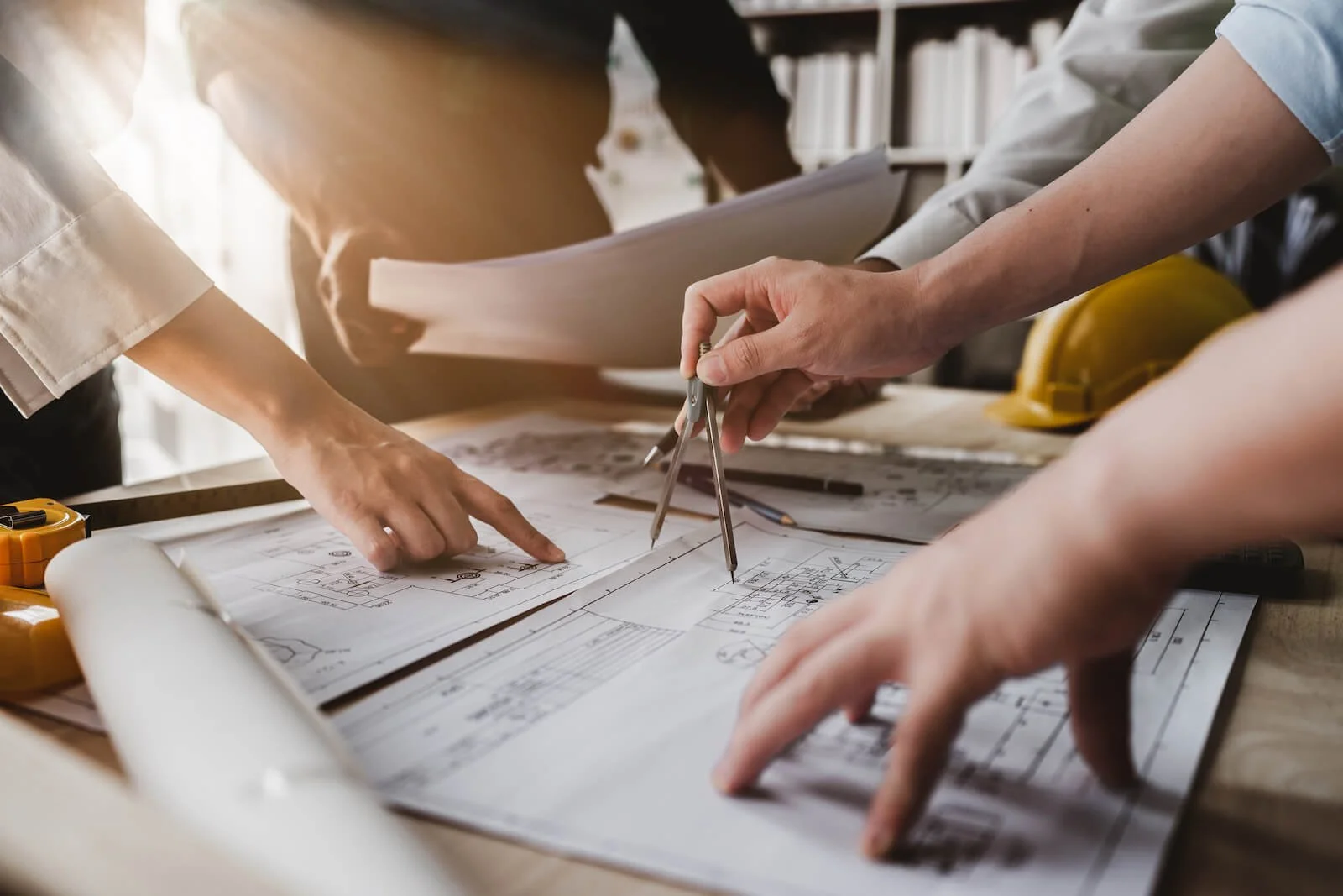 Multiple people working on architectural blueprints and drawings at a desk, with tools and safety helmets nearby.