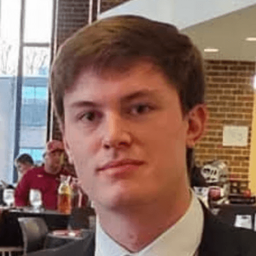 A young man with short brown hair wearing a white shirt and suit jacket, indoors at a dining area with brick walls and other people in the background.