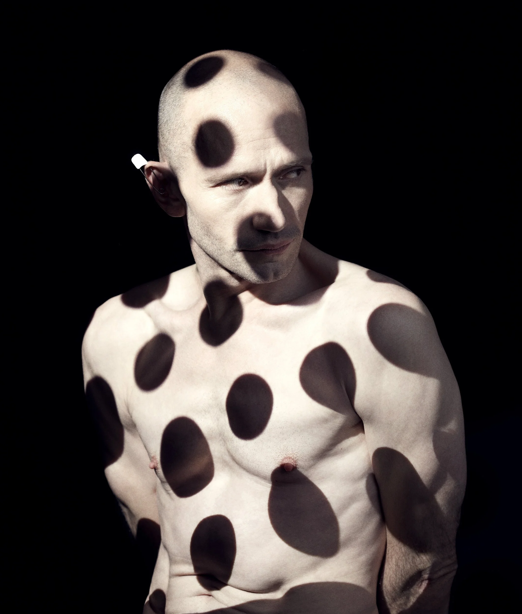 A shirtless man with a shaved head is seen against a dark background, with shadows cast across his face and body, creating an abstract pattern of large, dark spots.