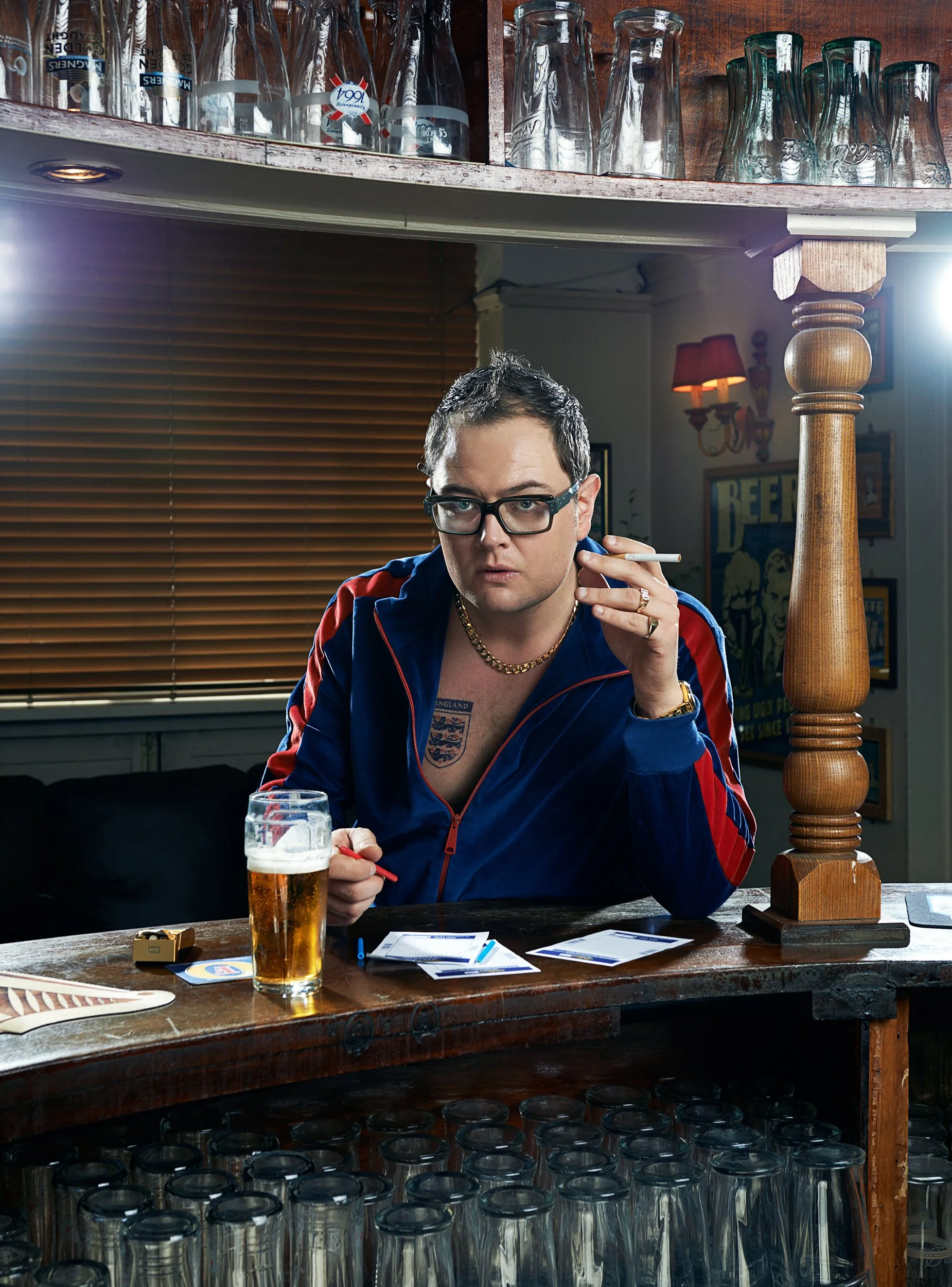 A man with glasses and a gold chain sitting at a bar with a beer in front of him, holding a cigarette, surrounded by glasses and bar items, with a background of wooden blinds and decorated walls.