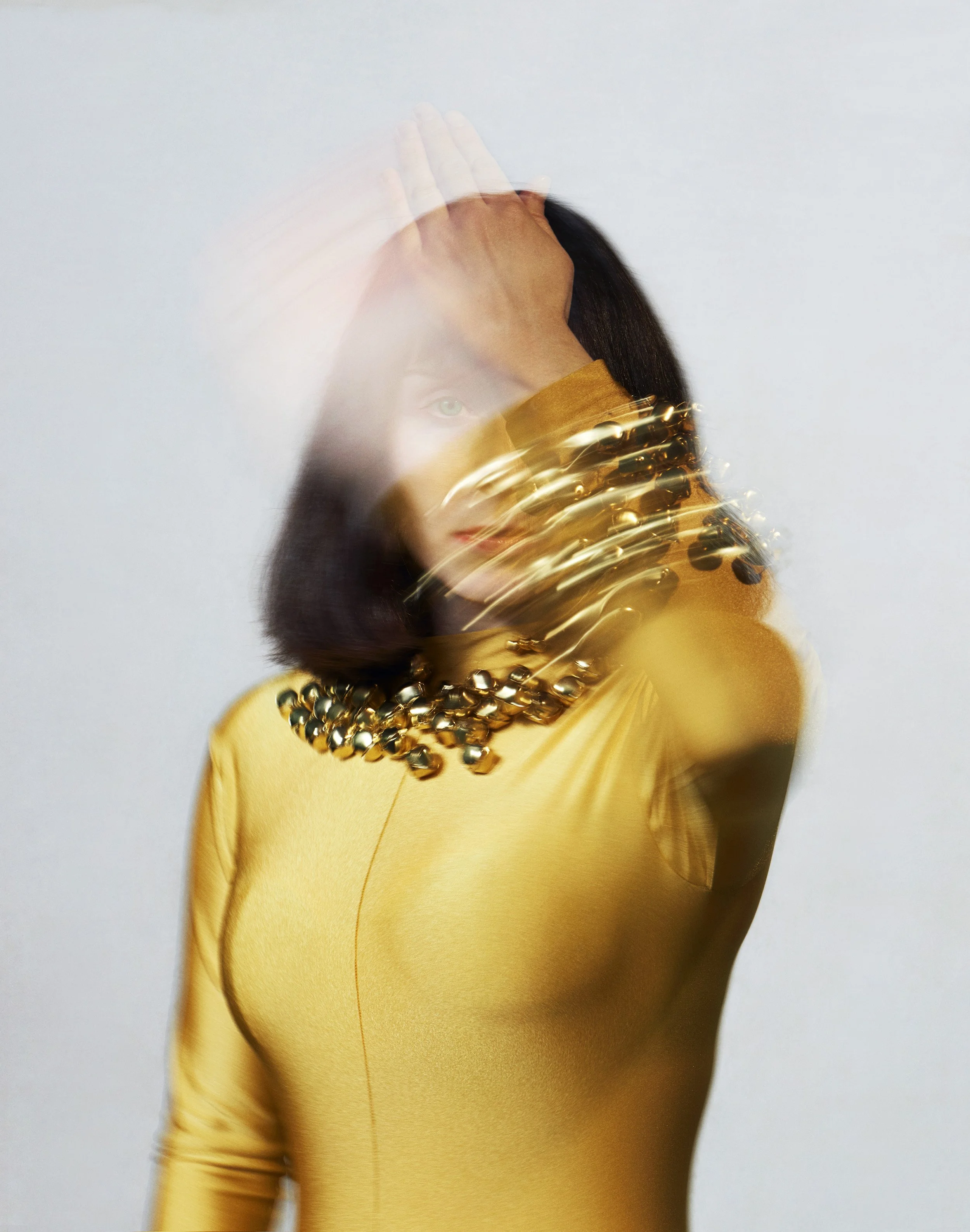 A woman in a yellow outfit with metallic jewelry covering her shoulder, forehead, and chest, with her hand on her head and her face partially obscured by motion blur.