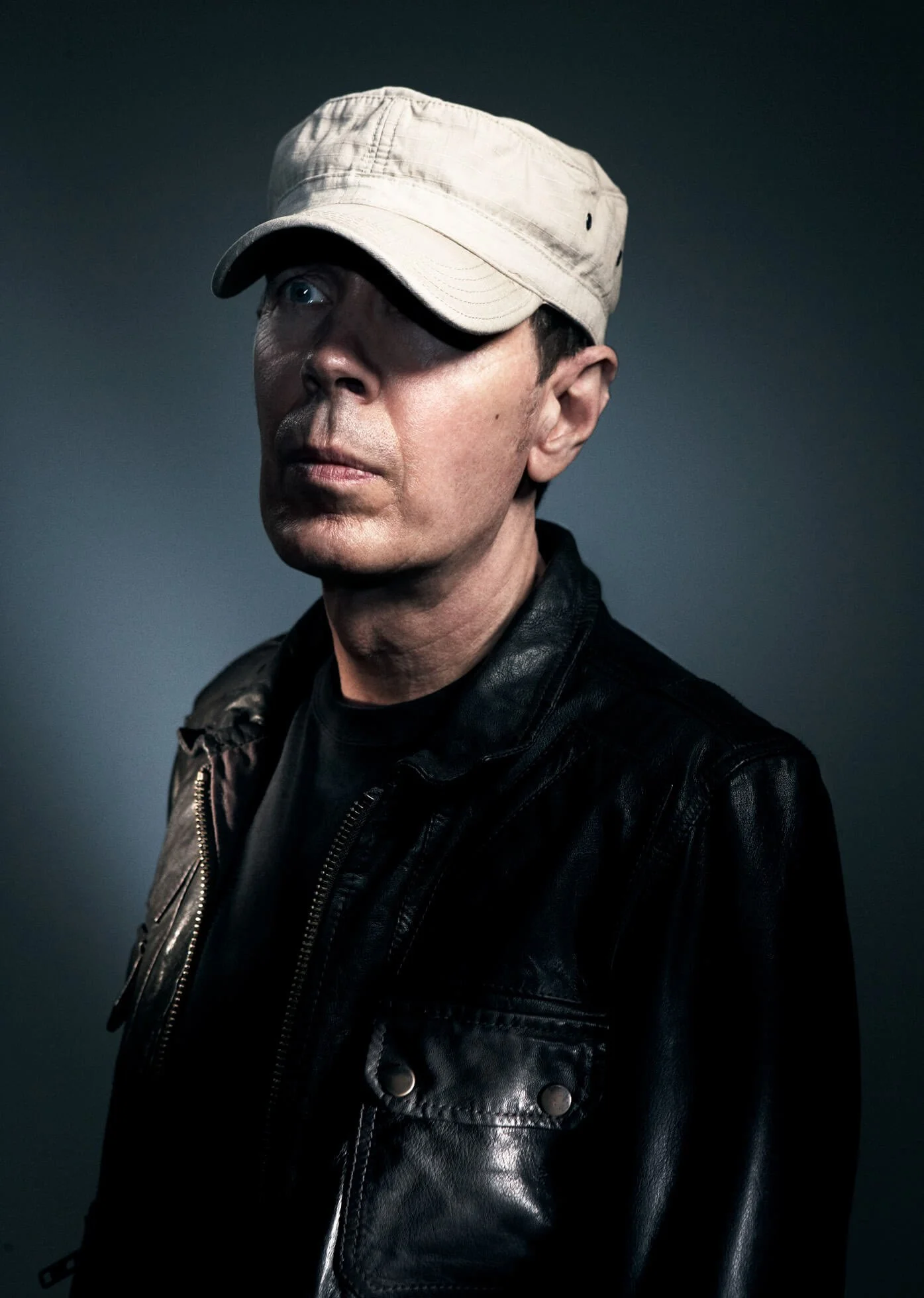 A man wearing a beige baseball cap and a black leather jacket, looking to the side against a dark background.