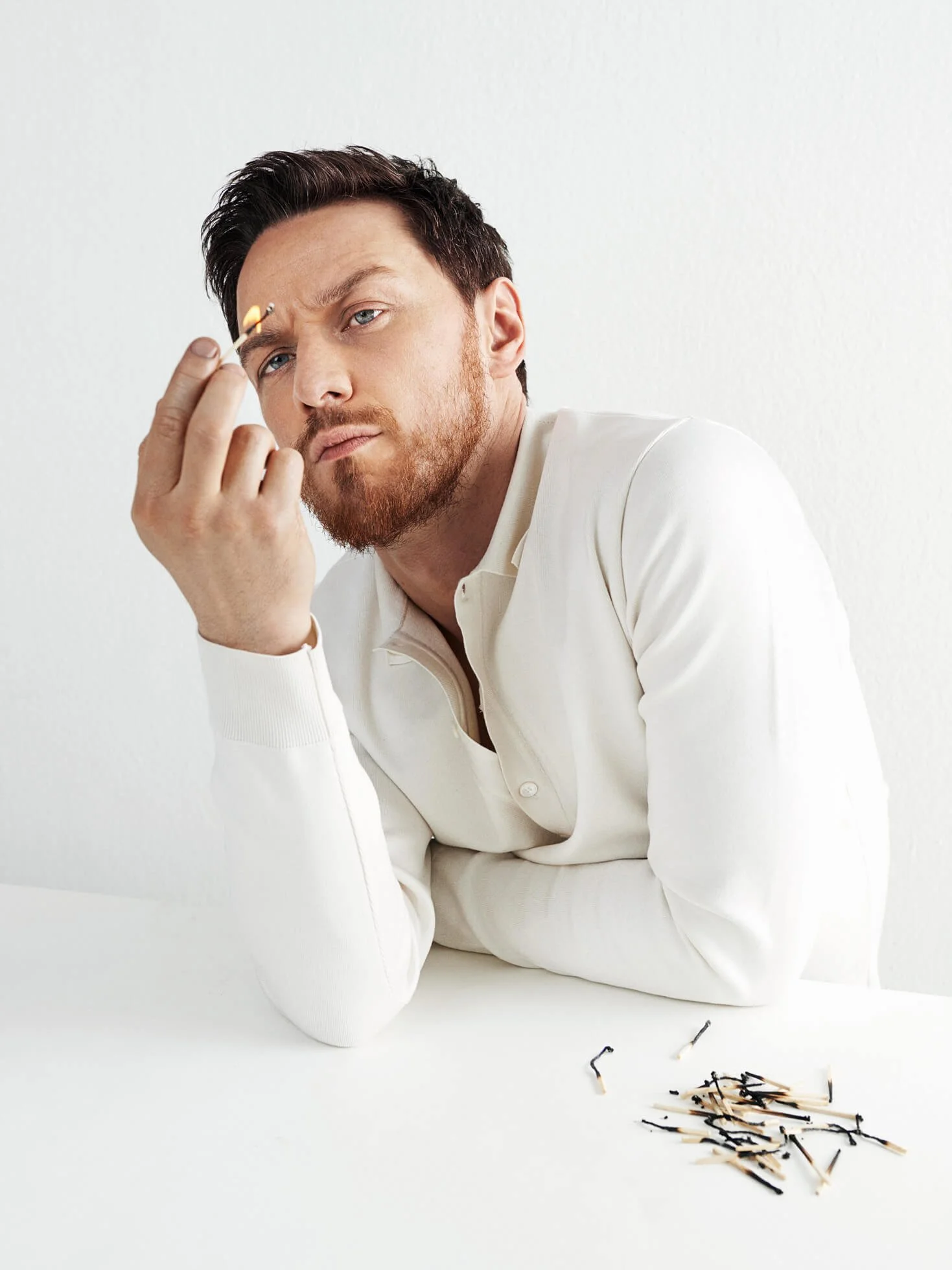 A man with blue eyes and ginger beard, wearing a white shirt, is leaning on a white surface, holding a lit matchstick. There are multiple burnt matches scattered on the surface.