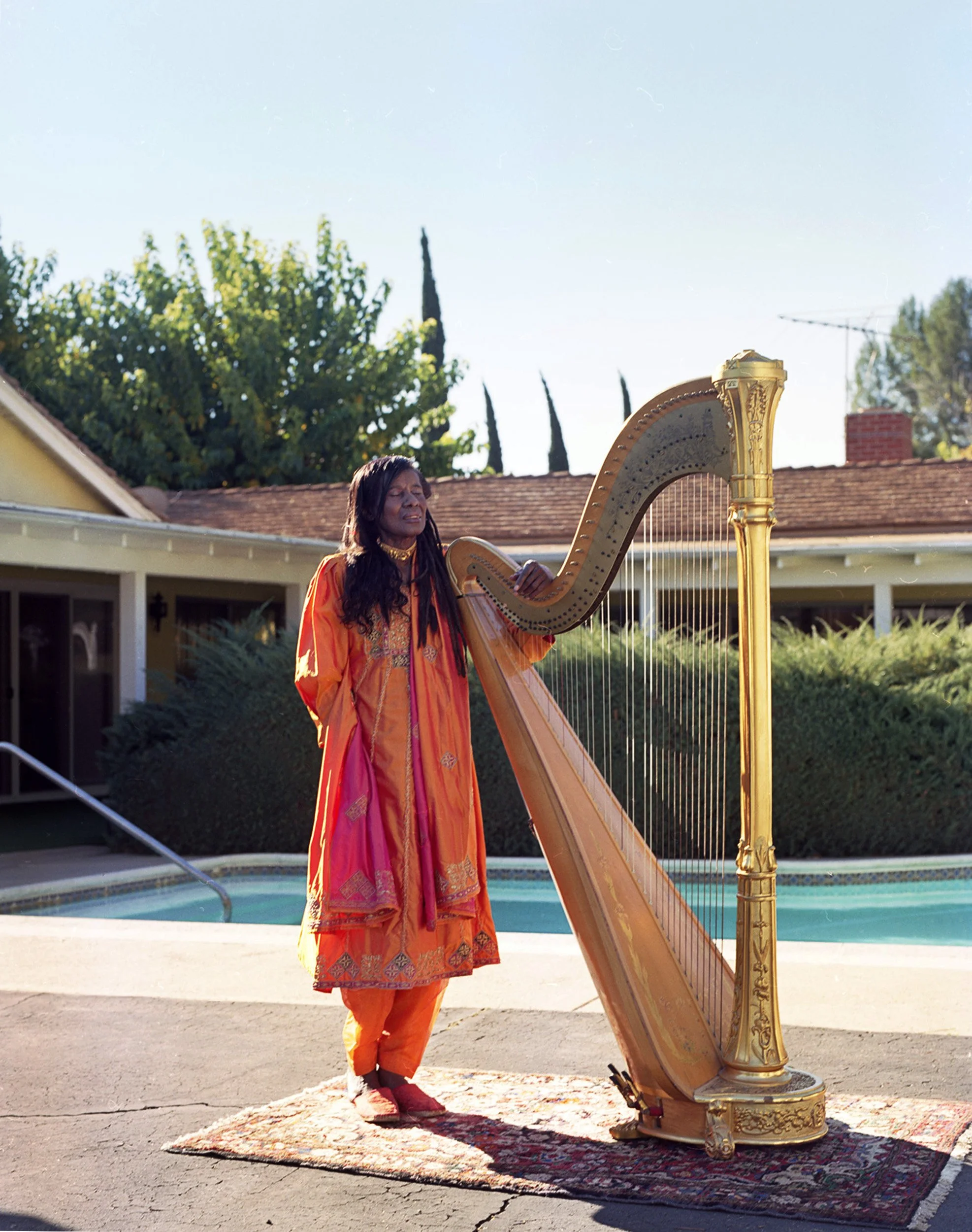 A woman dressed in colorful traditional clothing playing a golden harp outdoors on a rug, with a house, trees, and a pool in the background.
