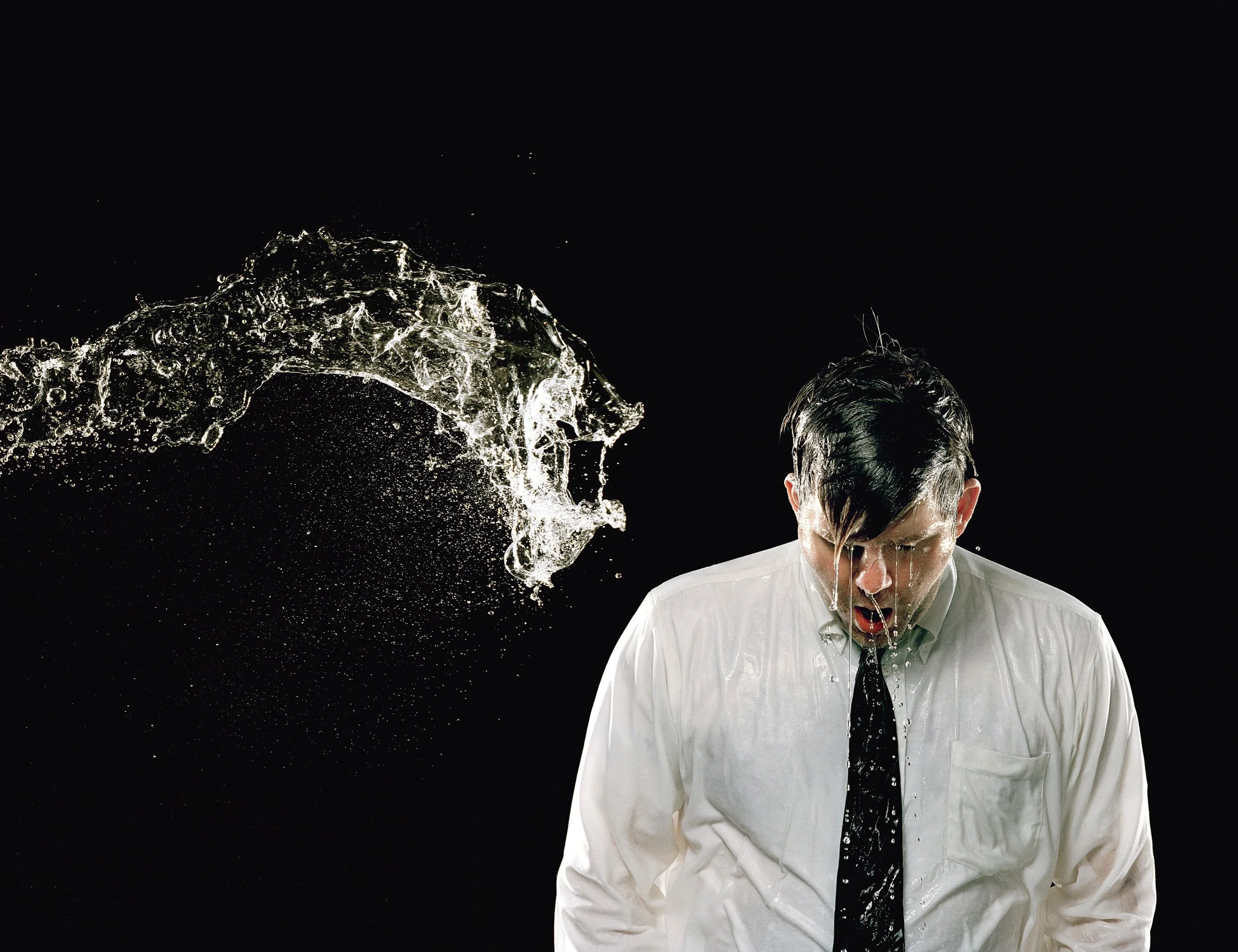 A man in a white shirt and black tie has water splashing on his face and head against a dark background.