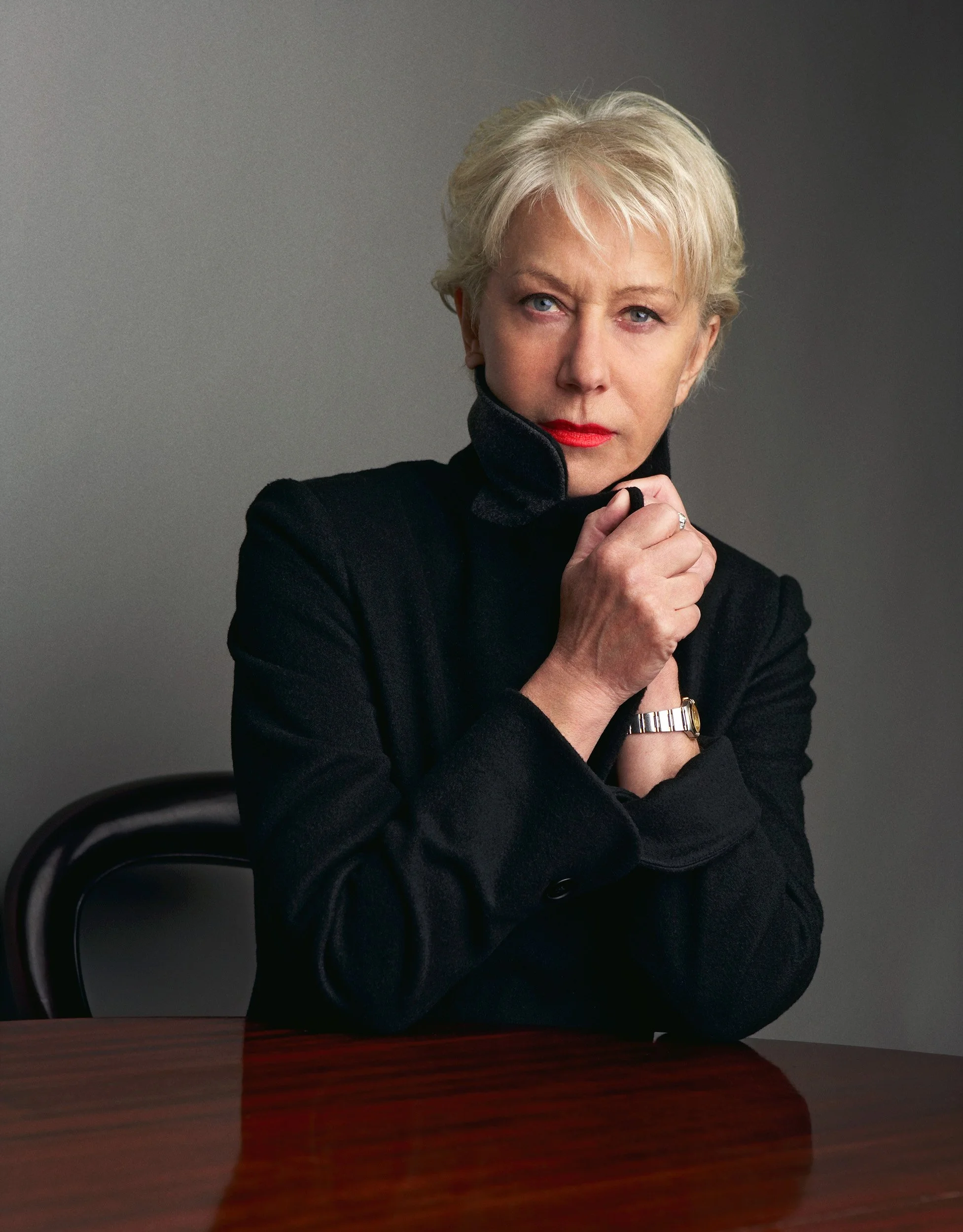 Close-up of a woman with short blonde hair, red lipstick, wearing a black jacket, sitting at a desk, holding her collar with her left hand, with a dark gradient background.