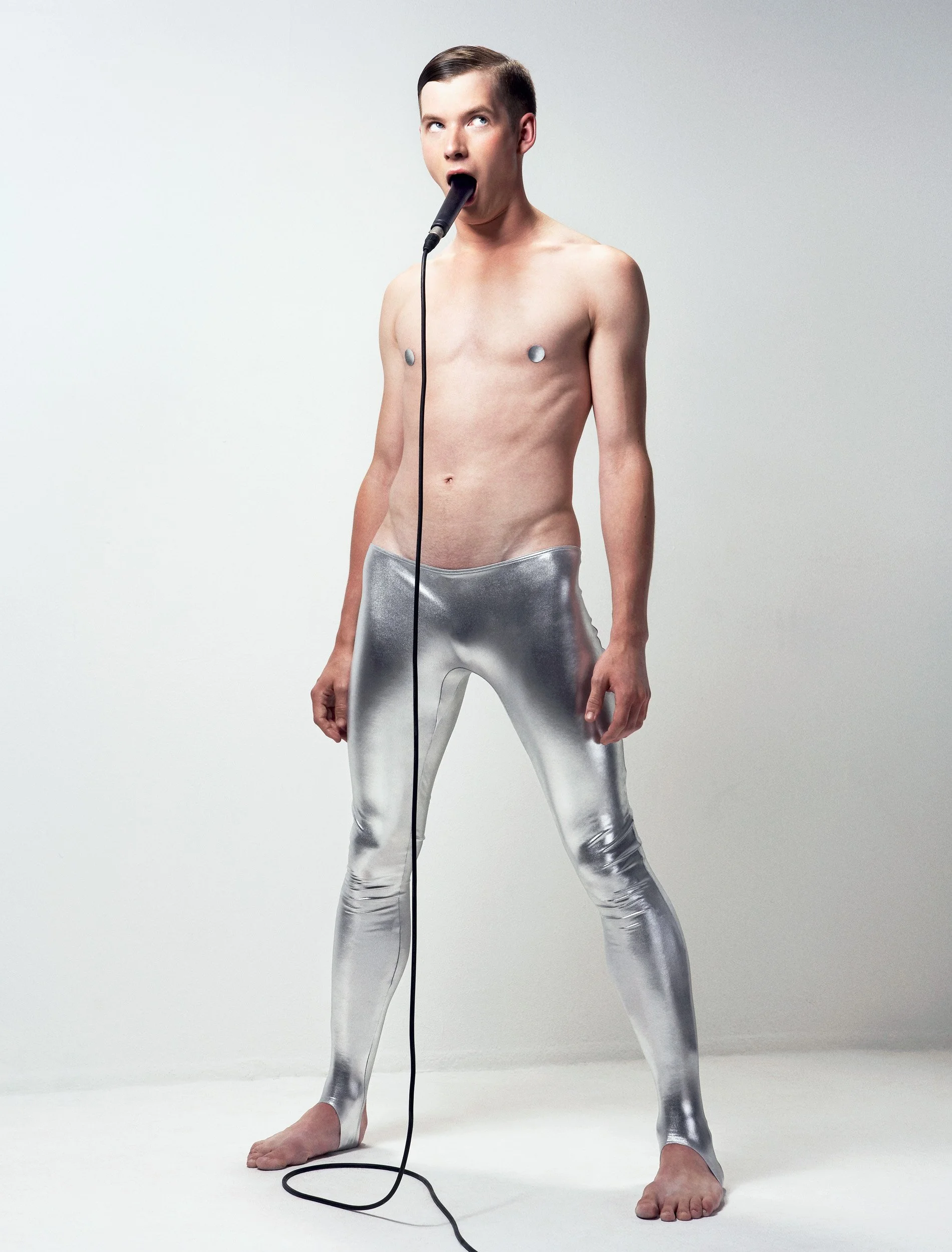 A person with short dark hair, wearing shiny silver pants, is standing against a plain white background, with metallic electrodes attached to their chest and skin. They have a microphone in their mouth and are holding a microphone cord. They appear t