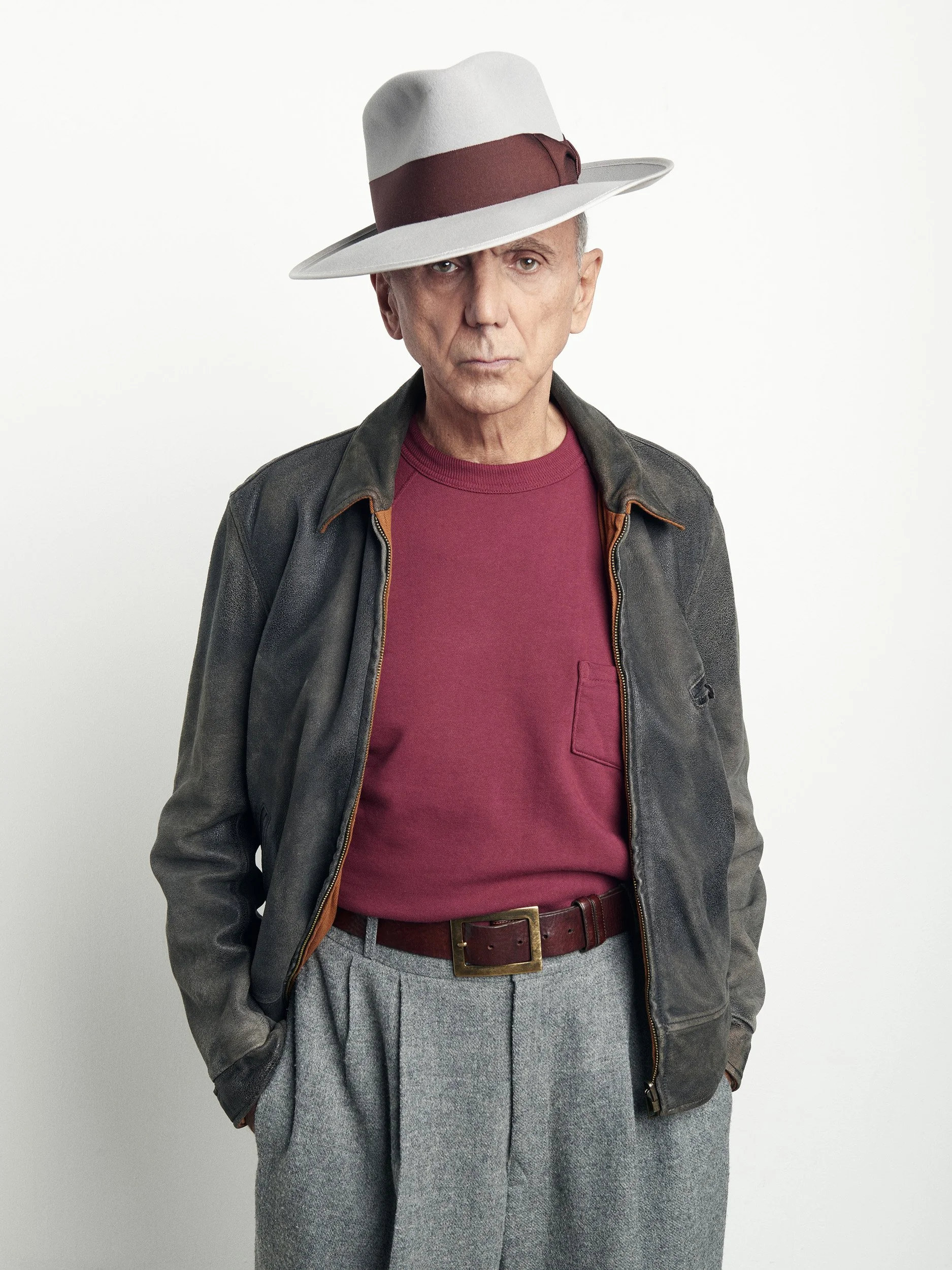 A man wearing a gray fedora hat with a maroon band, a gray leather jacket, a red T-shirt, and gray trousers.