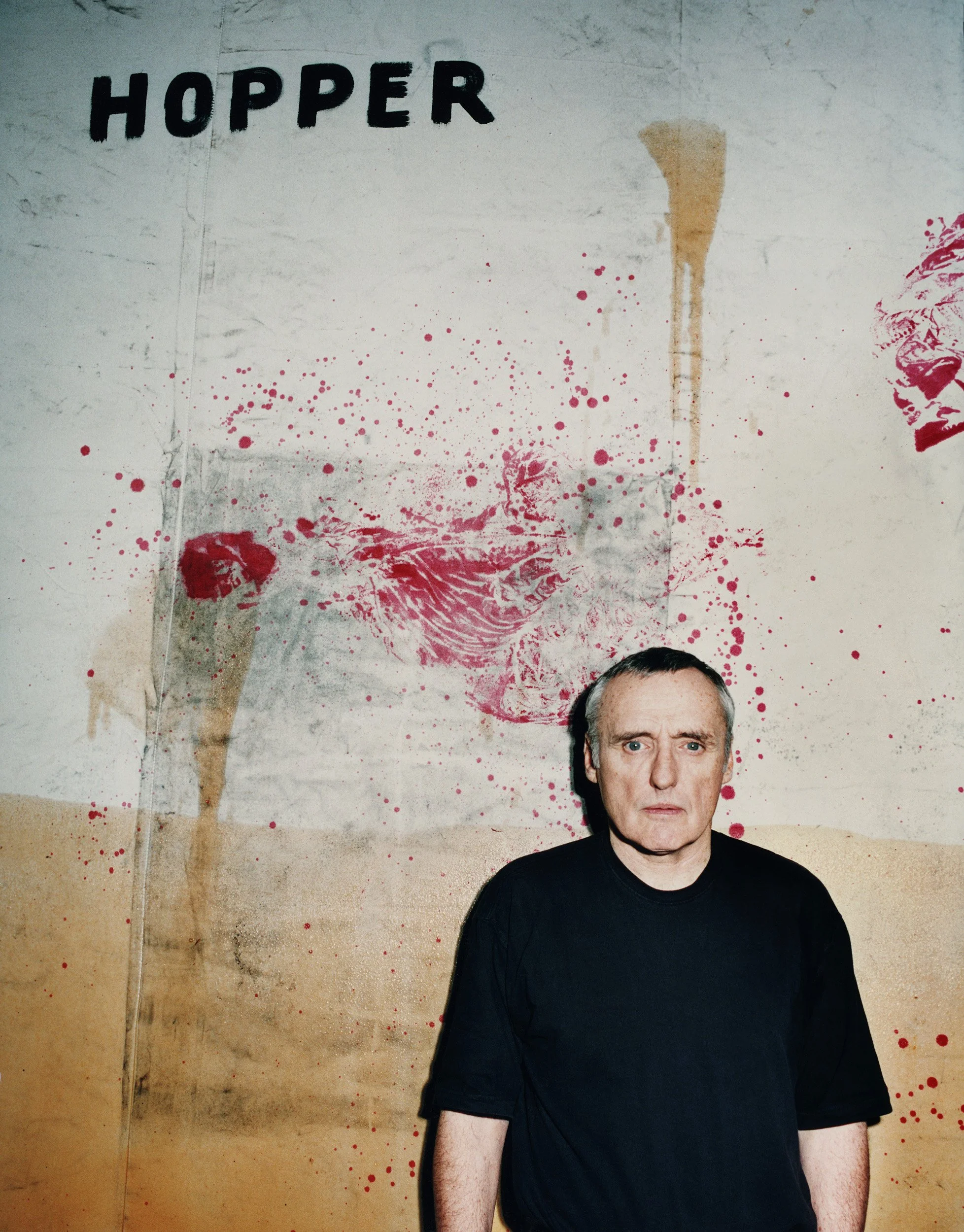 A person with light skin and gray hair wearing a black shirt standing in front of a wall with graffiti art of a bloody axe and a decapitated head in a splatter style, with the word 'HOPPER' written in bold black letters.