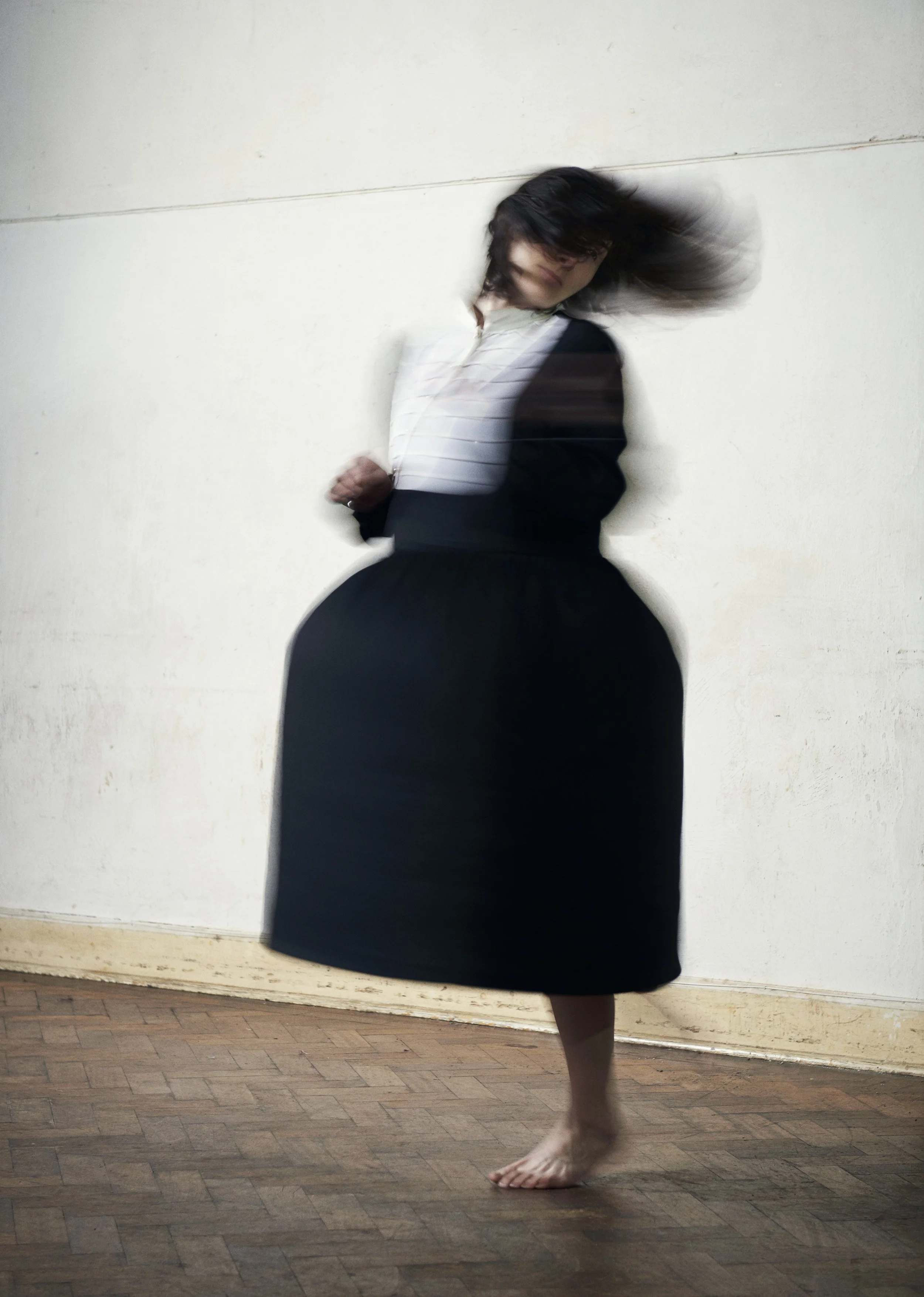 A woman with long dark hair leaning against a white wall, wearing a black skirt and a striped shirt, with one leg slightly raised and barefoot on wooden flooring.
