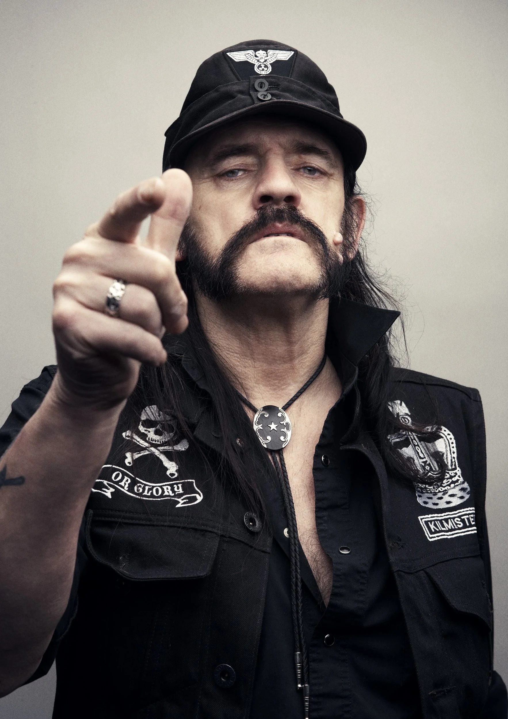 Man with long black hair and mustache, wearing a black police vest with patches, a necklace with a star and moon pendant, a ring, and a black cap with a white eagle insignia, pointing towards the camera.