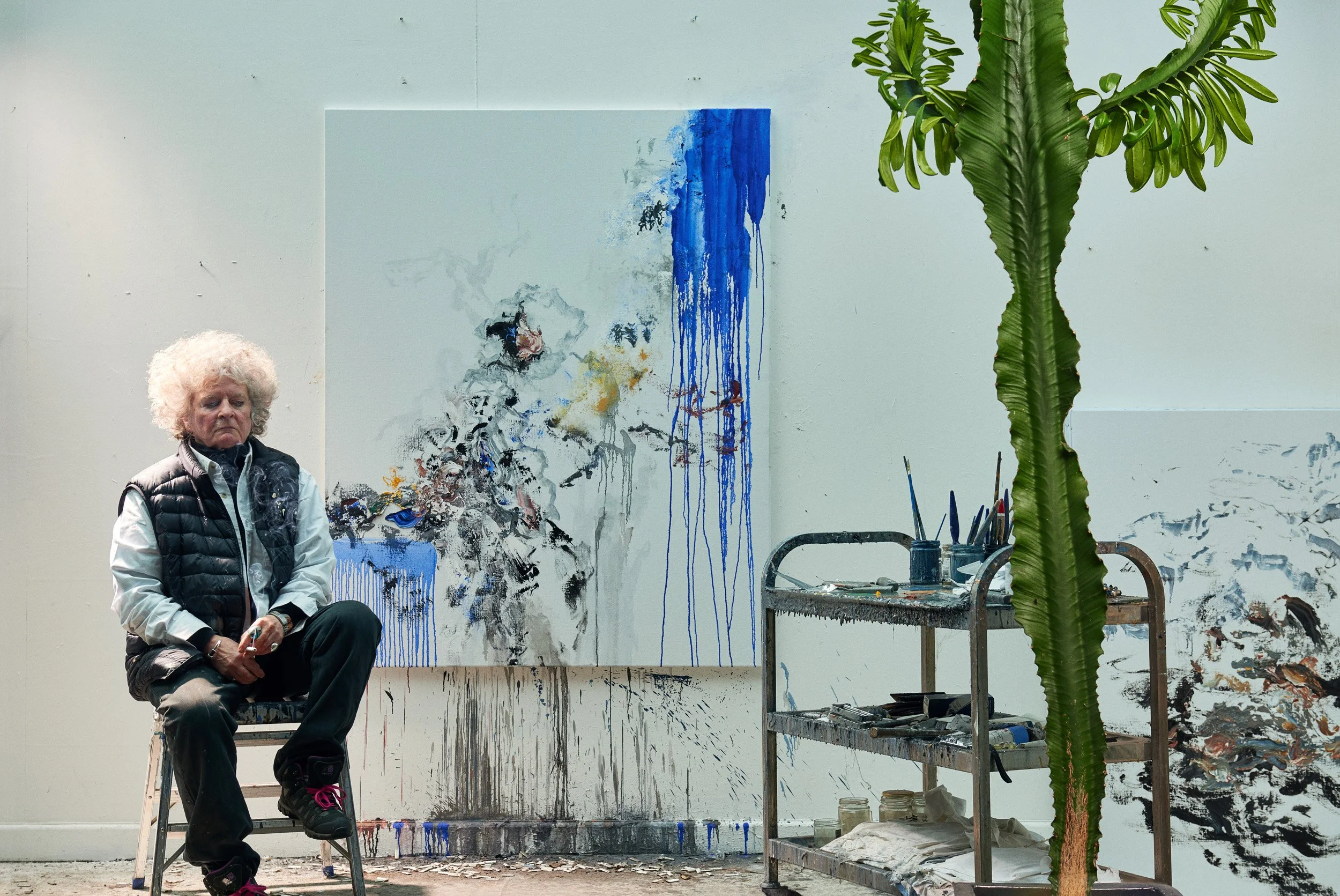 An elderly woman with curly white hair sitting on a step stool in an art studio, with a large abstract painting behind her featuring blue, black, and yellow paint splatters and drips. There is a tall green plant with large leaves in the foreground, a