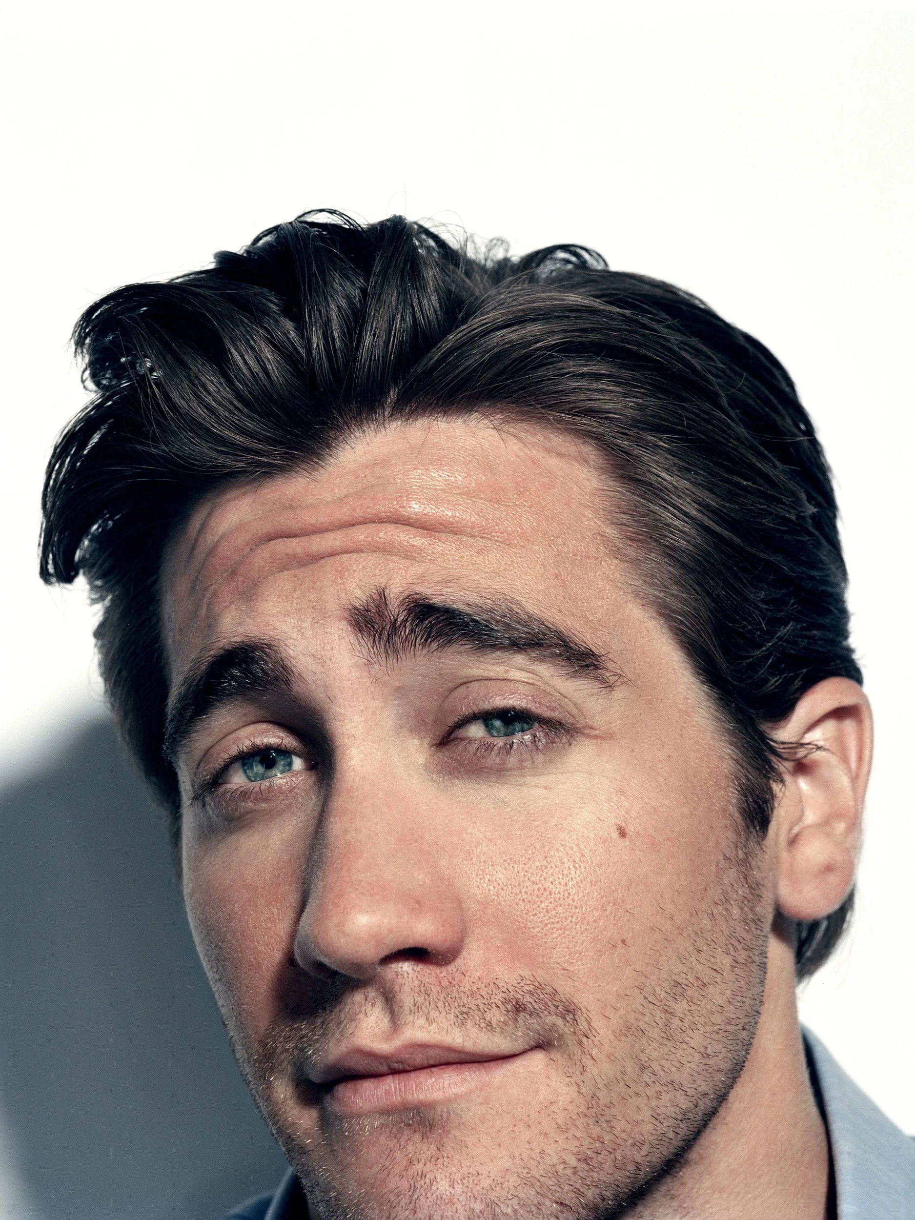 Close-up of a man's face showing black hair, blue eyes, and facial stubble, with part of his forehead and cheek visible.