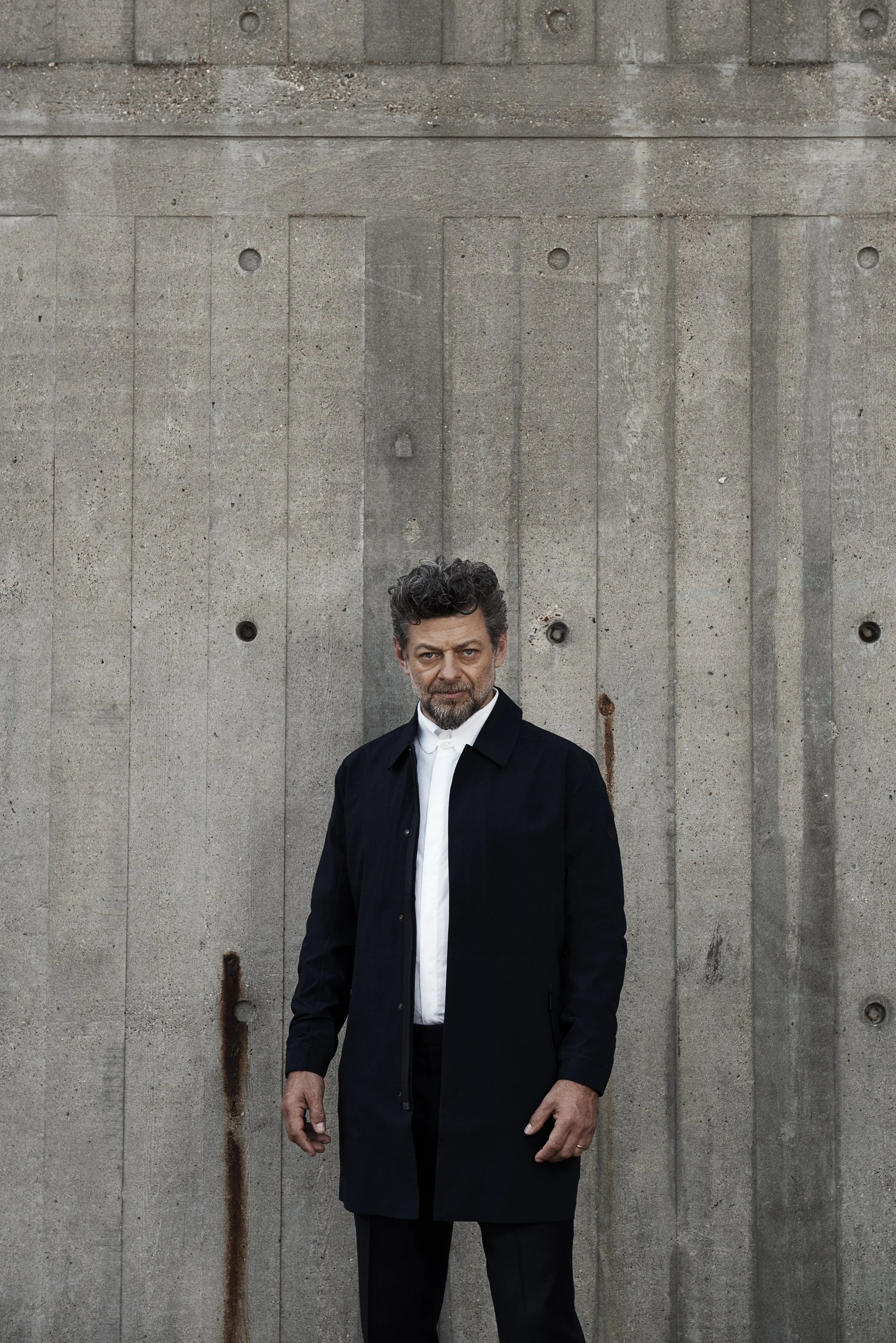 A man with curly dark hair and a beard wearing a long dark coat over a white shirt standing outdoors in front of a weathered concrete wall with visible rust stains and metal fittings.