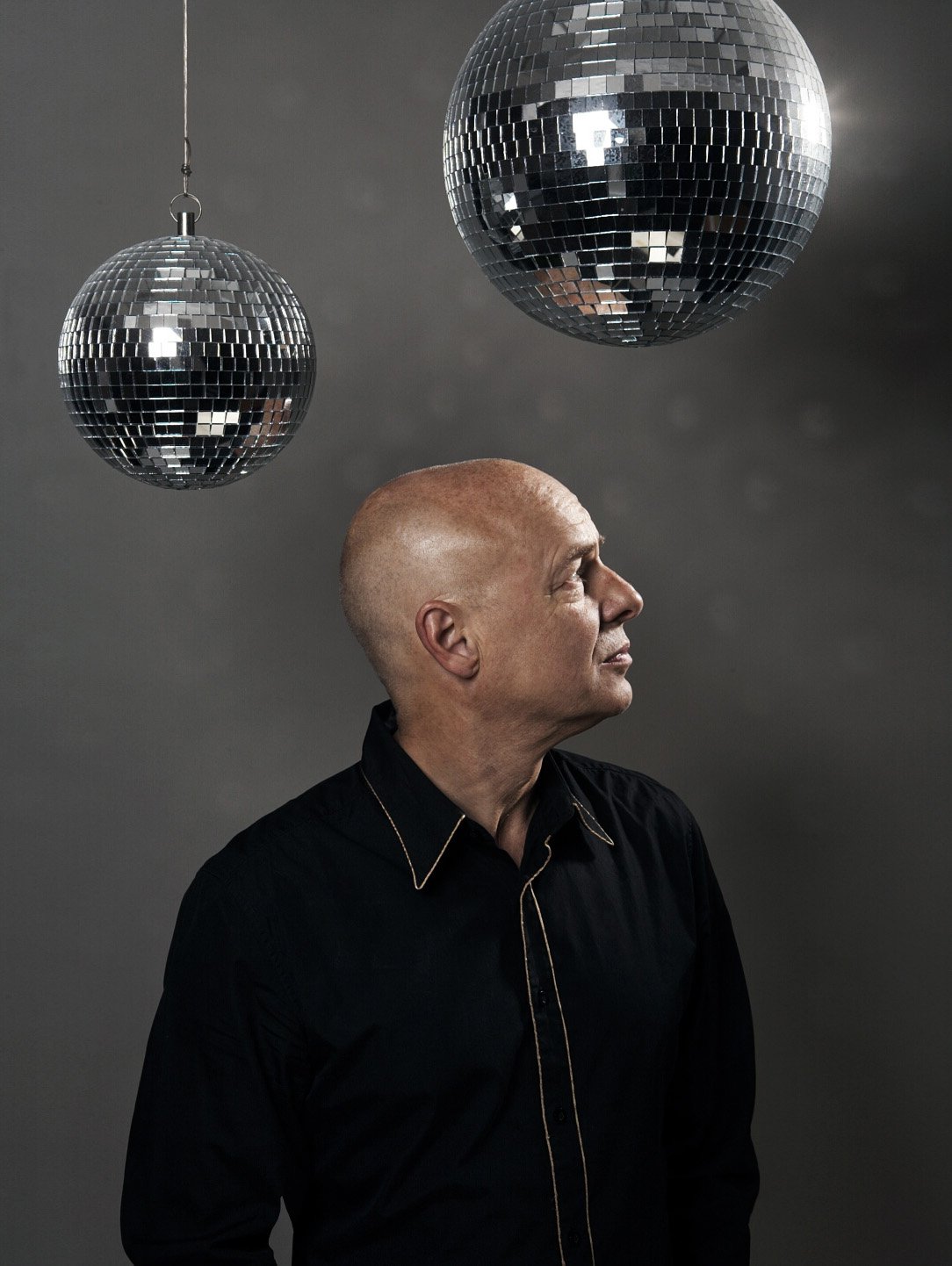 A bald man with light skin wearing a black shirt with gold trim staring at two hanging disco balls against a gray background.