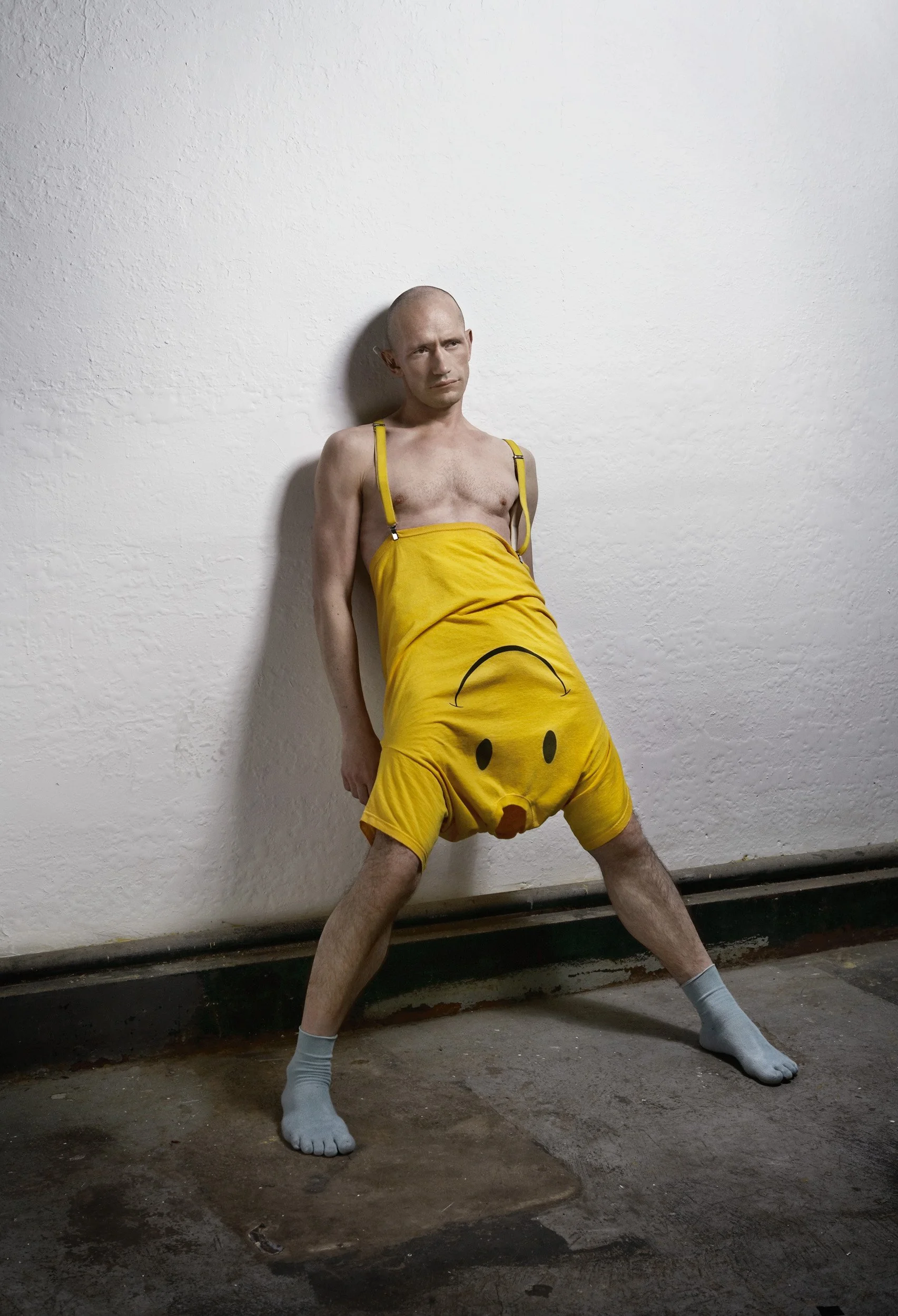 A man with a shaved head and thin build wearing a yellow smiley face patterned outfit with suspender straps and white socks, standing against a white wall with a serious expression.